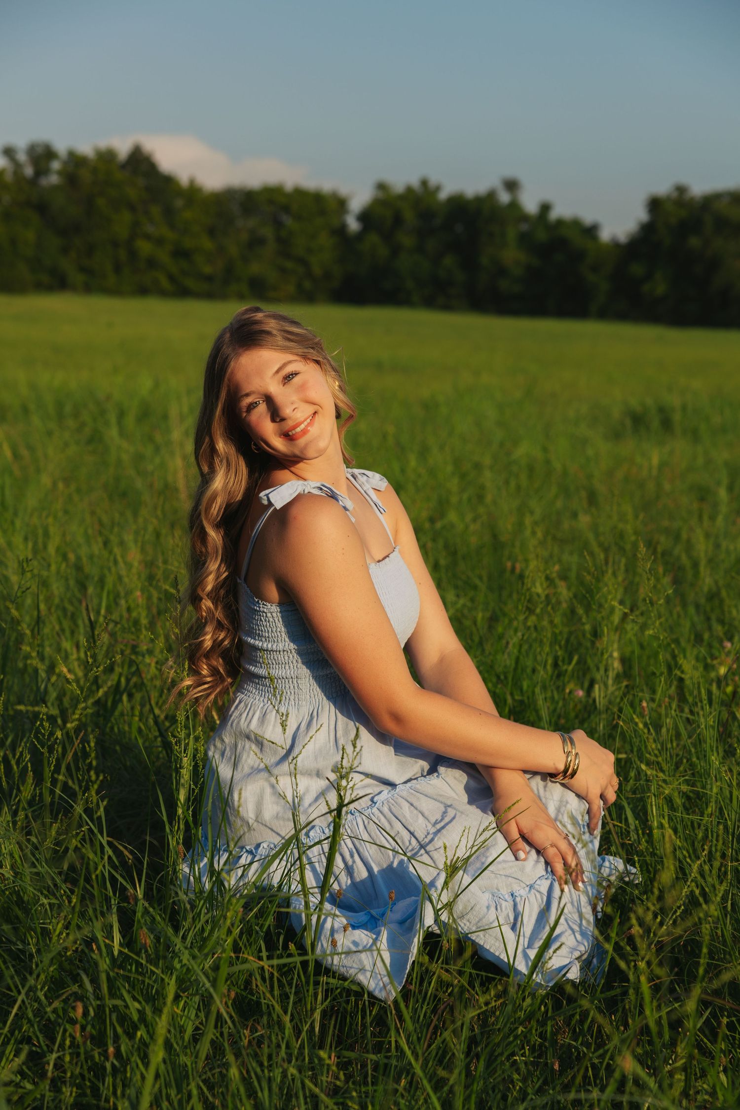 Smiling young woman in white floral dress sitting in tall green grass on a sunny summer day.