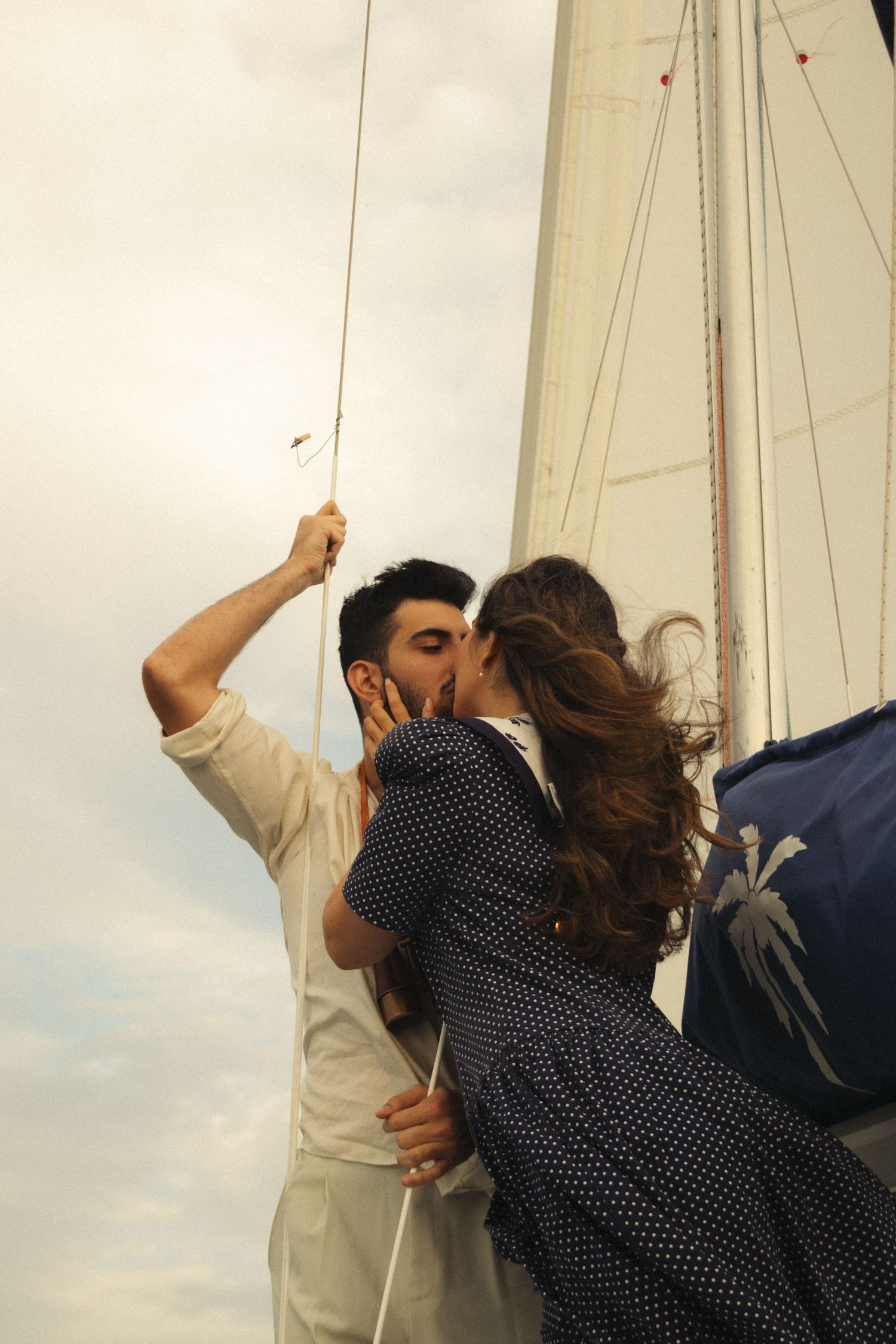 A couple shares a romantic kiss on a sailboat with white sails against a bright glowing sky.