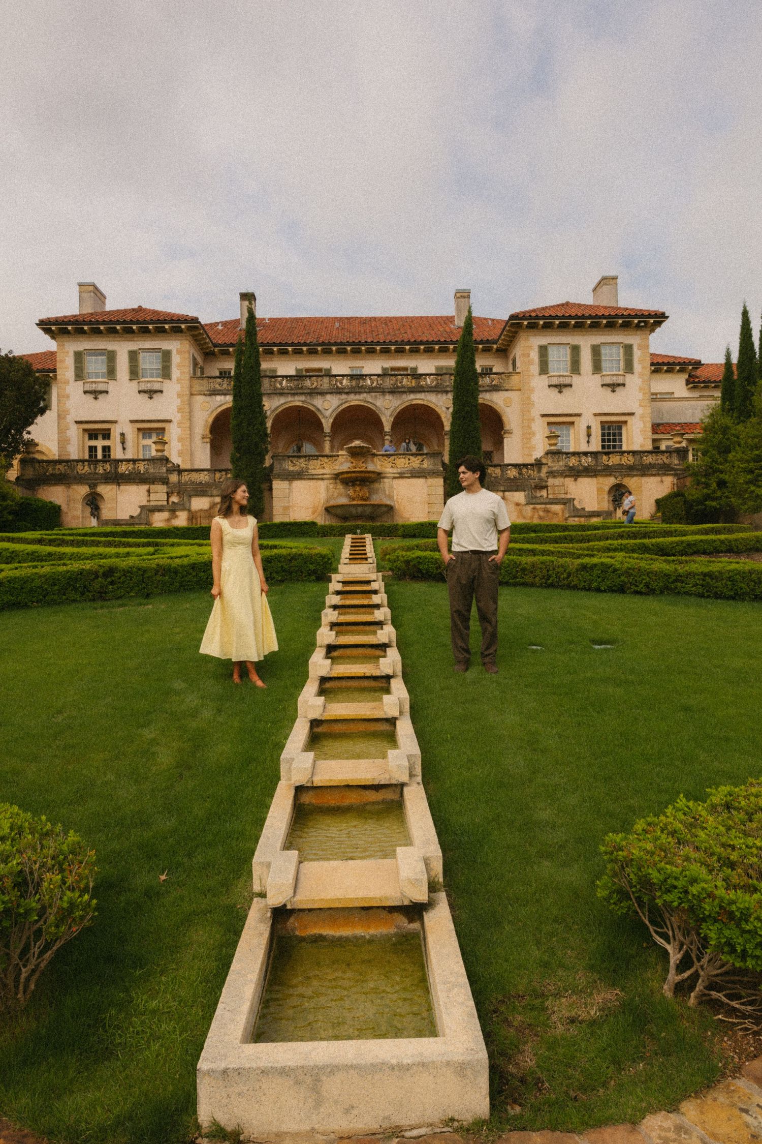 Couple standing beside ornate water feature at elegant Italian-style mansion garden.