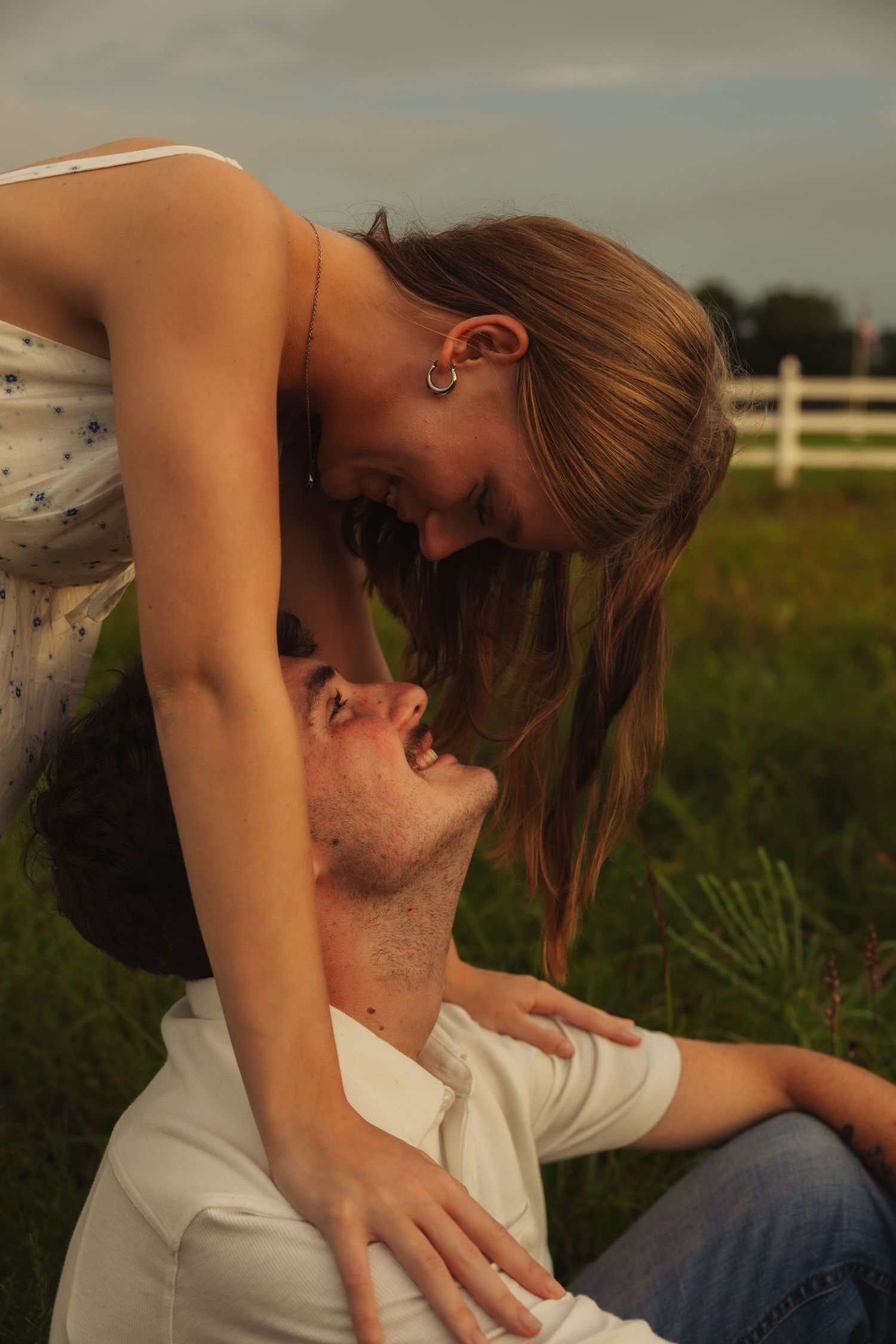 Romantic couple in a field, woman leaning over smiling man near white fence at golden hour.