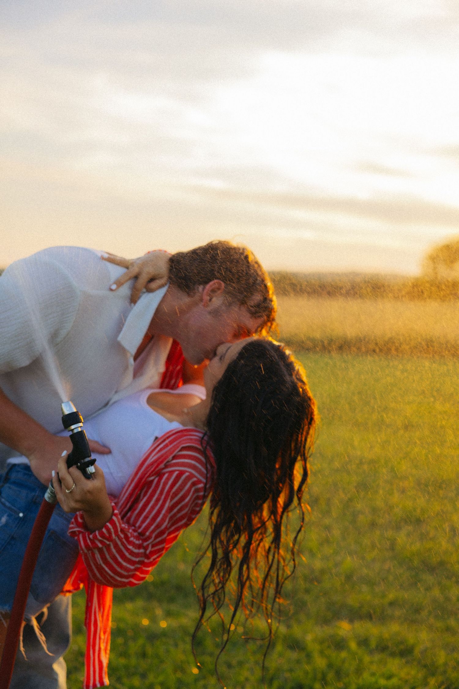 Couple sharing a romantic dip kiss outdoors at golden hour while holding a garden hose in a green field.