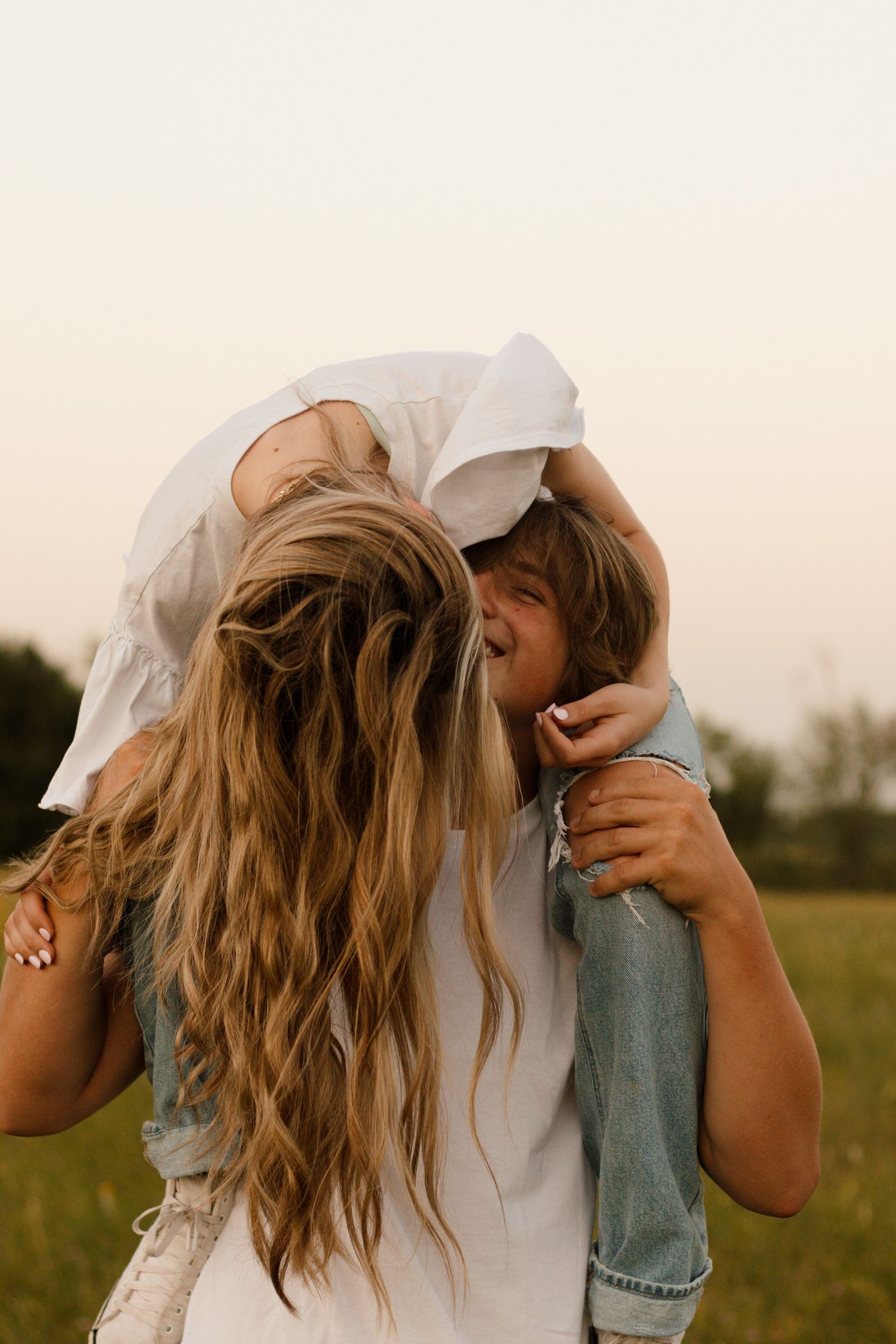 Laughing couple embracing playfully in a sunlit field, woman with long blonde hair leaning back.