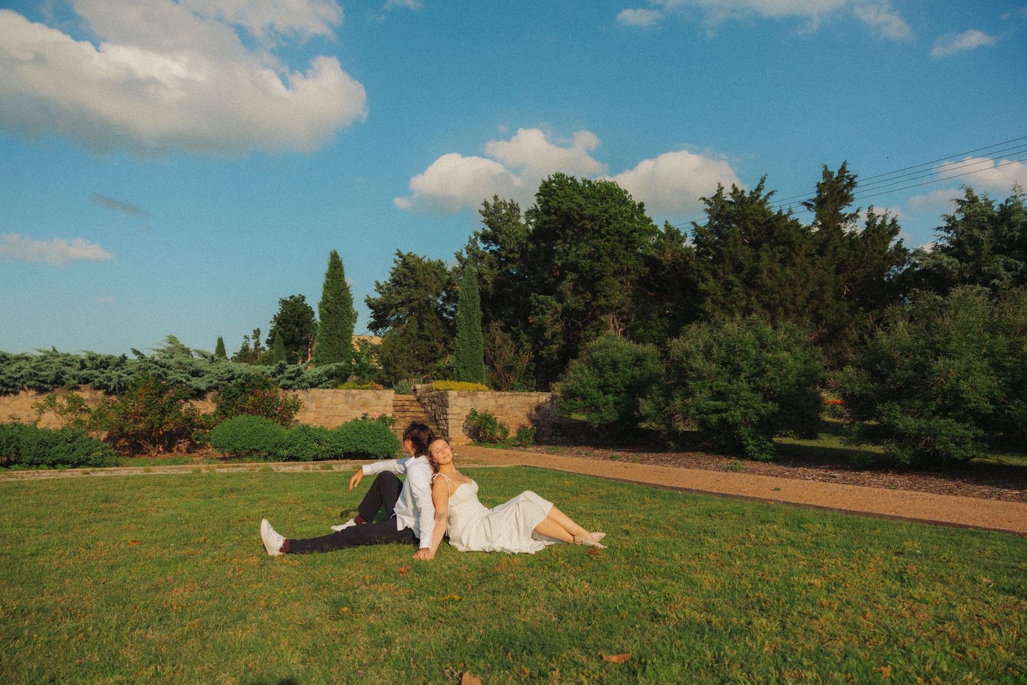 Couple in wedding attire sitting together on green grass with trees and blue sky backdrop.