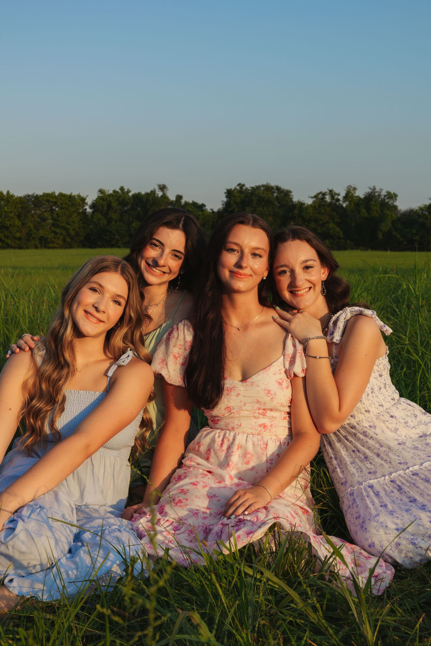 Four smiling young women in floral dresses sitting together in a green field at sunset.