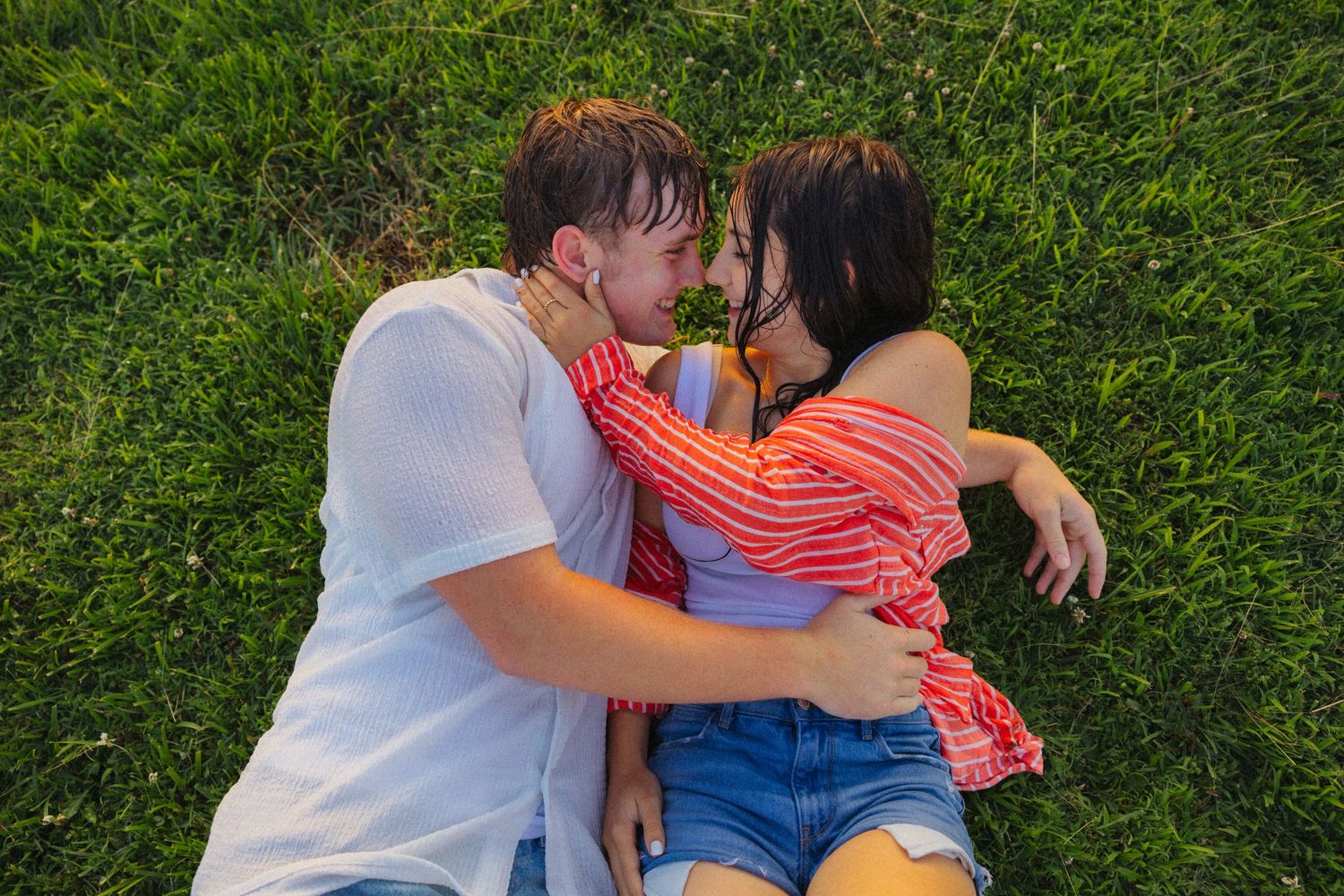 Young couple lying together on green grass, facing each other in a tender and intimate moment.