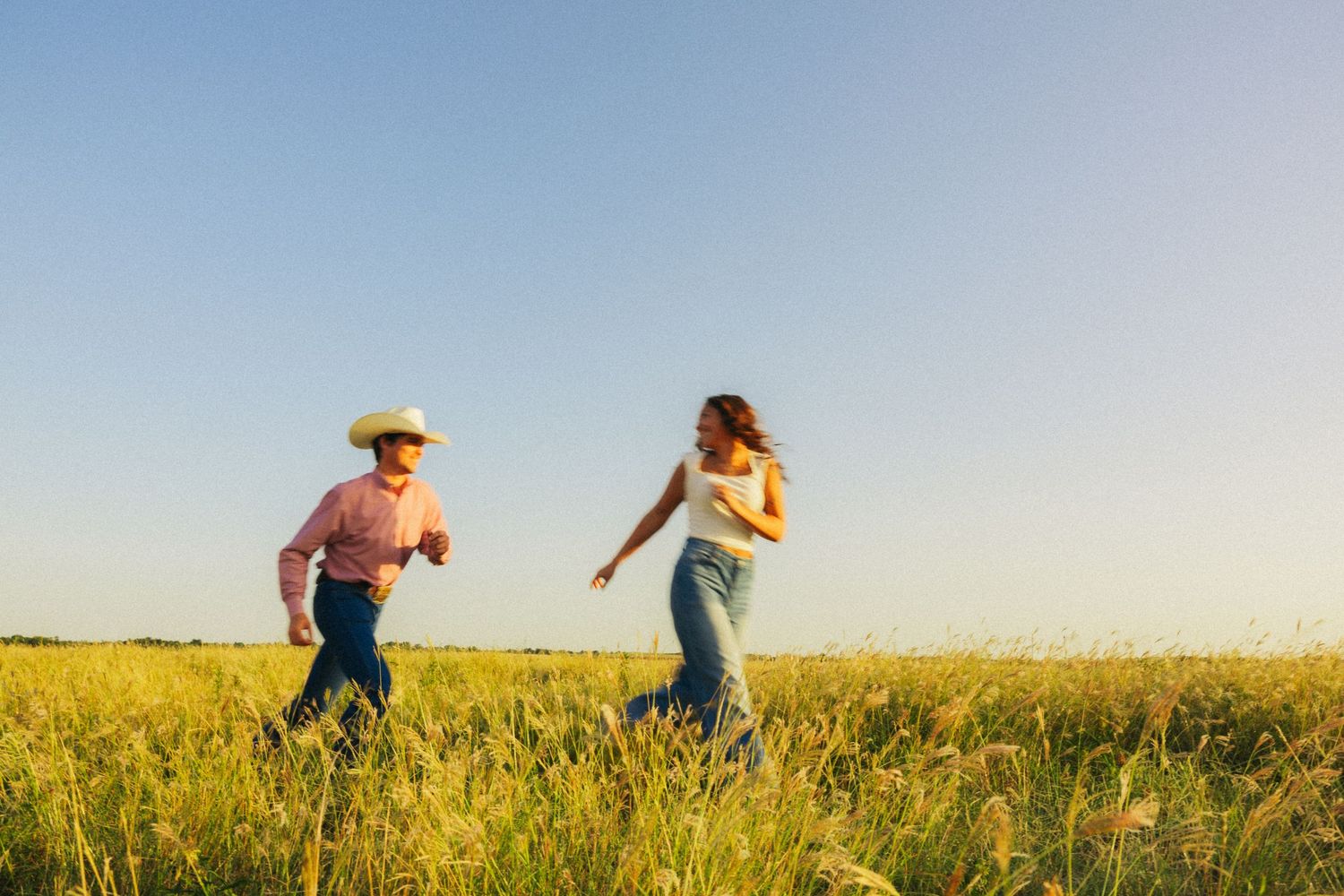 A couple runs freely through a golden grass field under a bright blue sky, with the man wearing a cowboy hat.