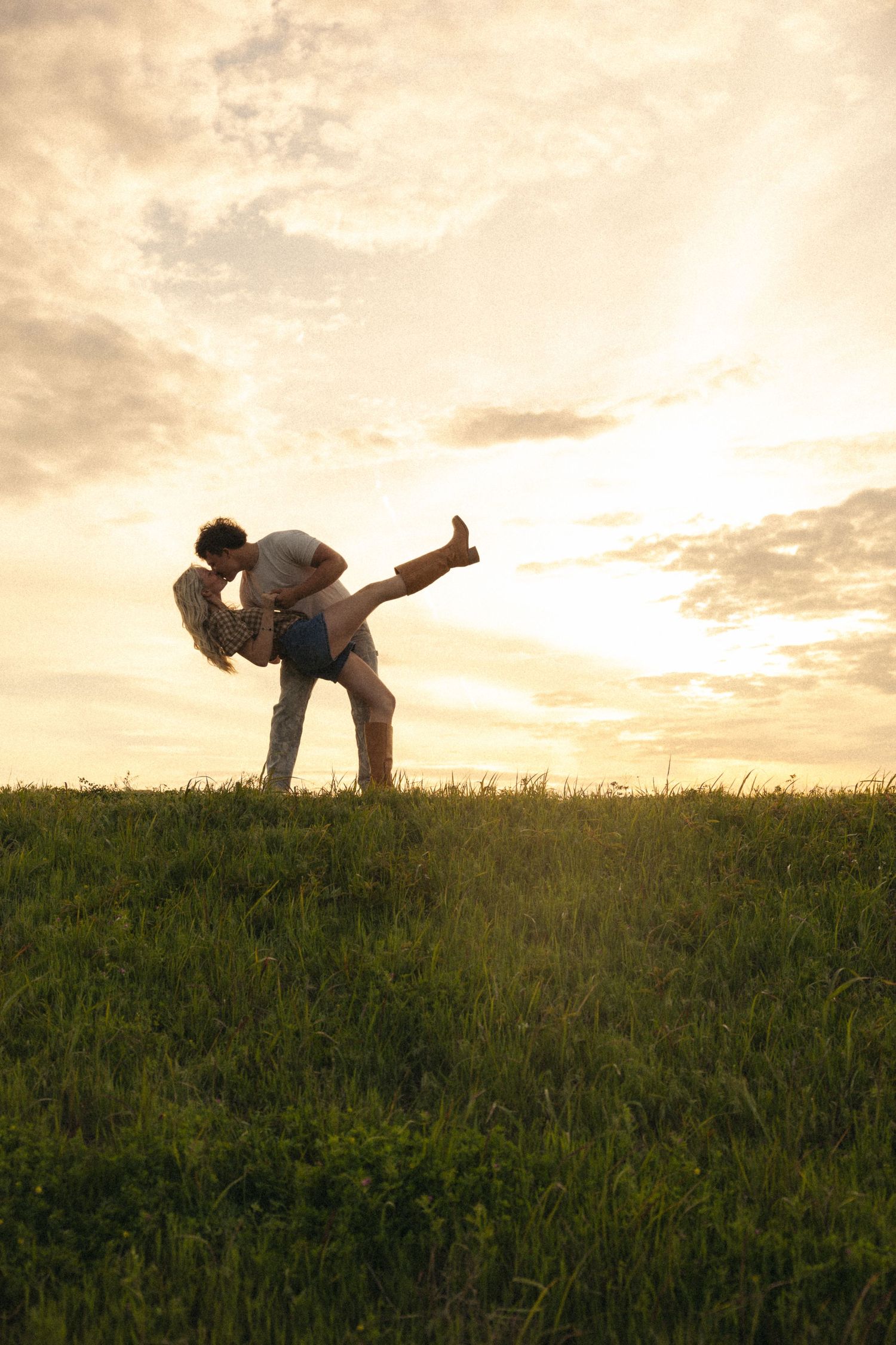 Romantic couple silhouetted against golden sunset sky on grassy hillside.