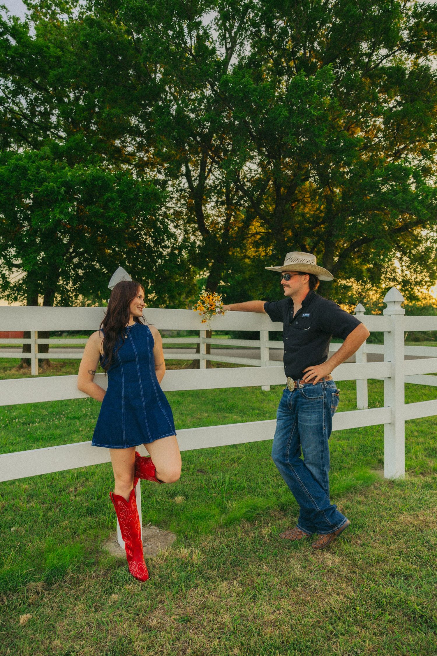 Couple laughing together by a white fence at sunset, woman in red cowboy boots and blue denim dress.