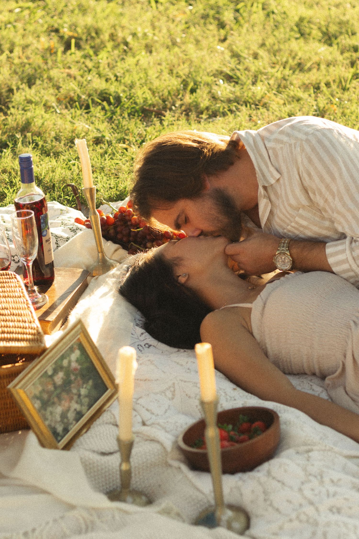 Romantic couple kissing during an outdoor picnic with candles, wine, and strawberries on a blanket at sunset.