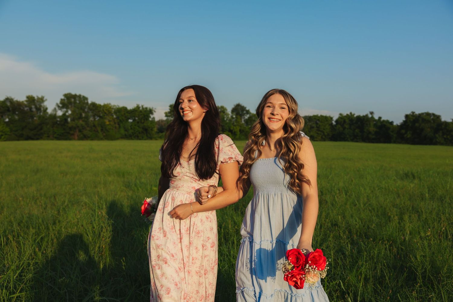 Two smiling young women in floral and blue dresses holding red bouquets in a sunny green meadow.