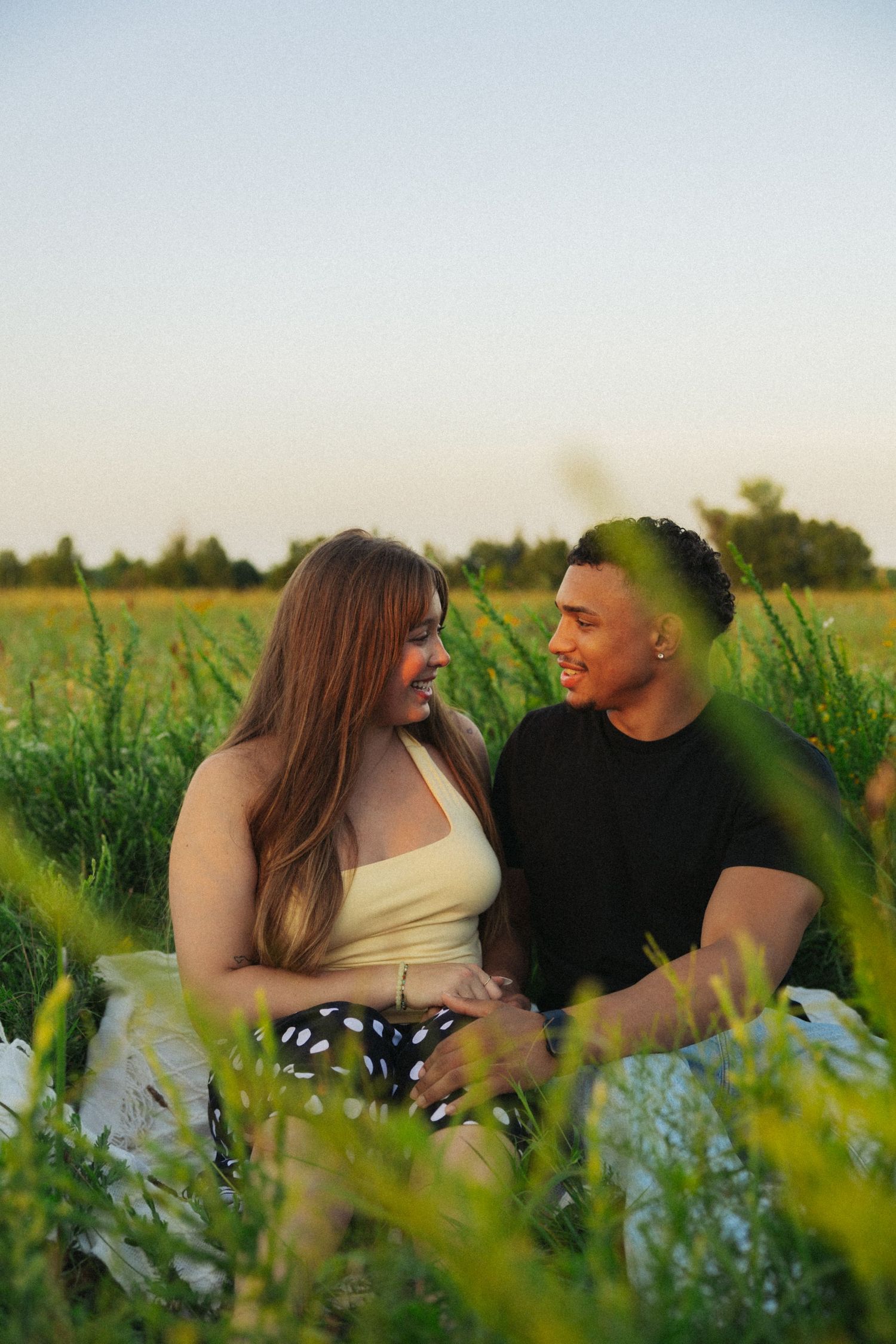 A happy interracial couple sits together in a lush green field during golden hour, smiling at each other romantically.
