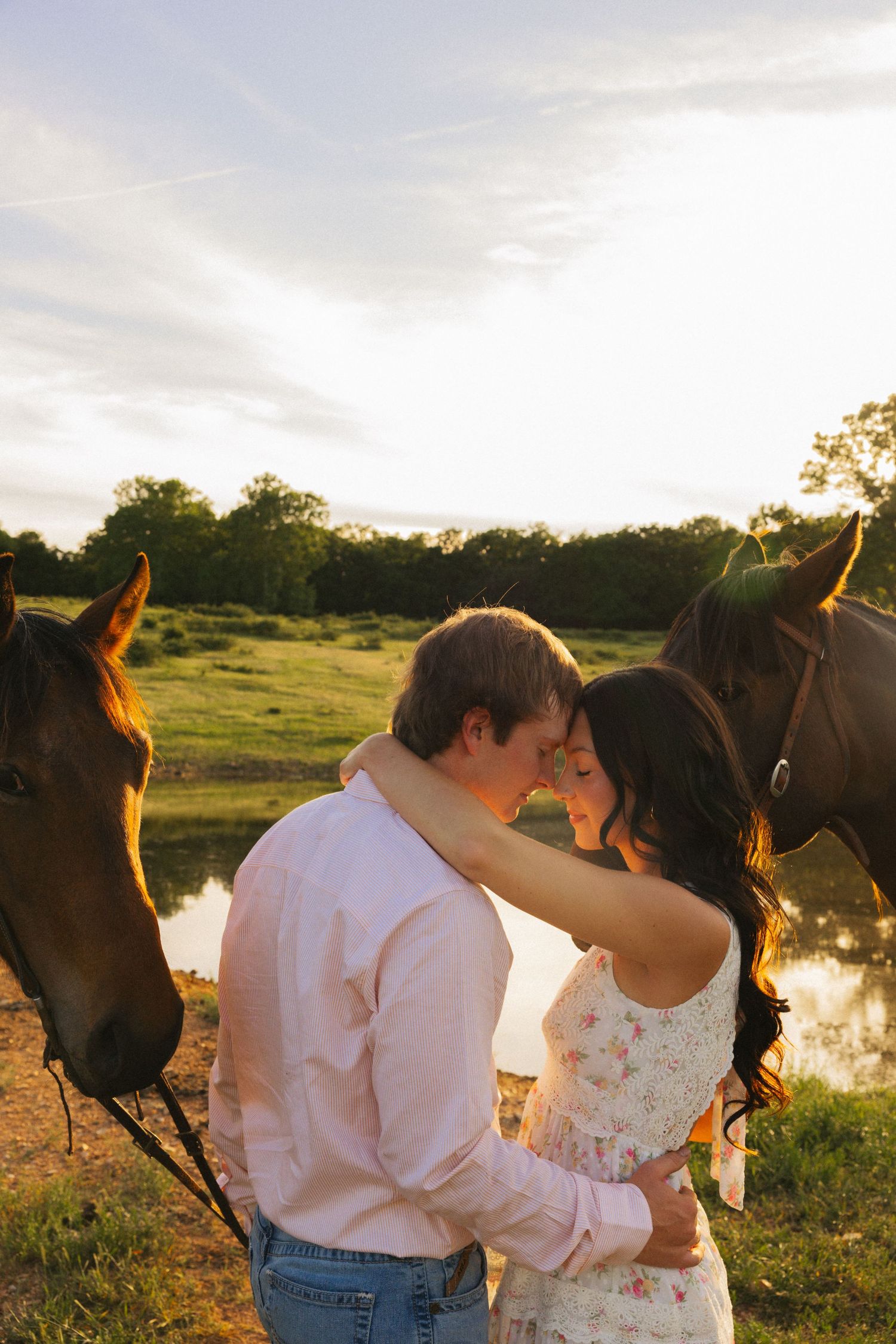 Couple sharing tender moment between two horses at golden sunset by a pond.