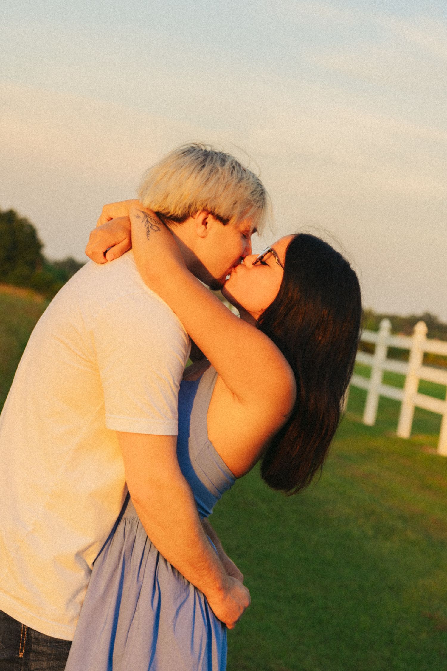 A couple shares a romantic kiss at golden hour near a white fence, the woman in a blue dress embracing warmly.