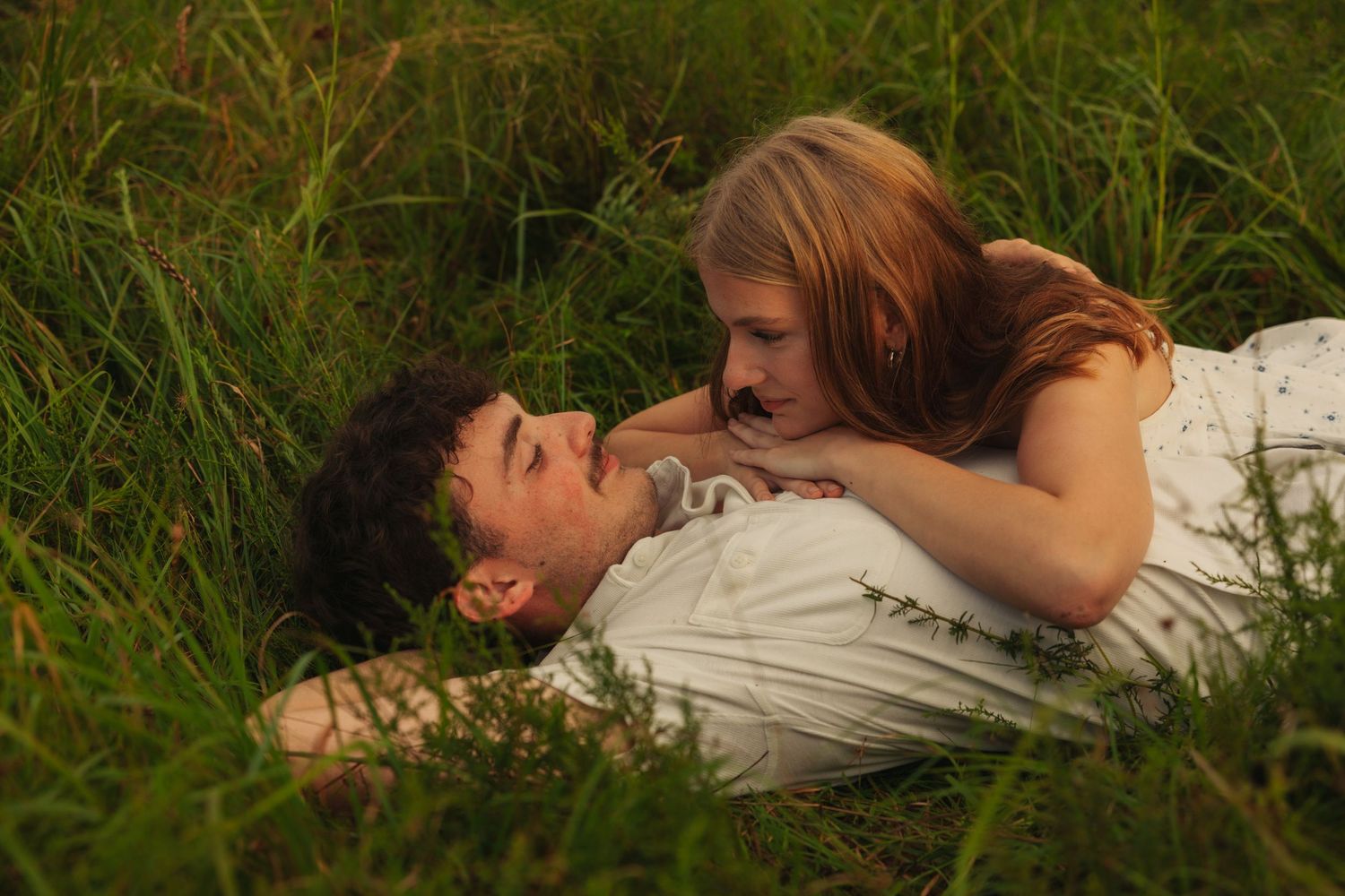 Romantic couple lying in green grass field, woman leaning over man smiling in warm golden light.
