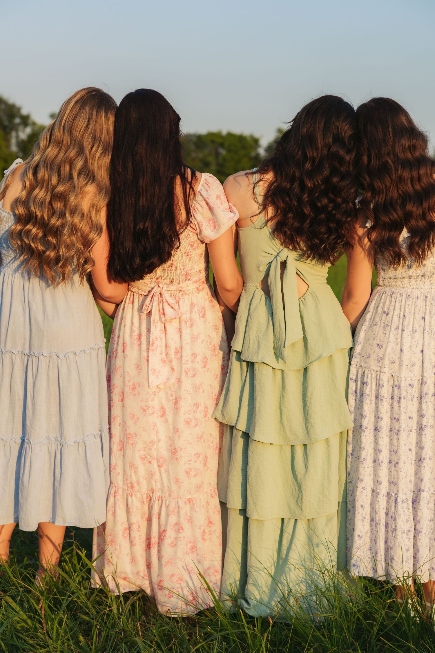 Four women in pastel summer dresses standing together from behind in a sunny green field.
