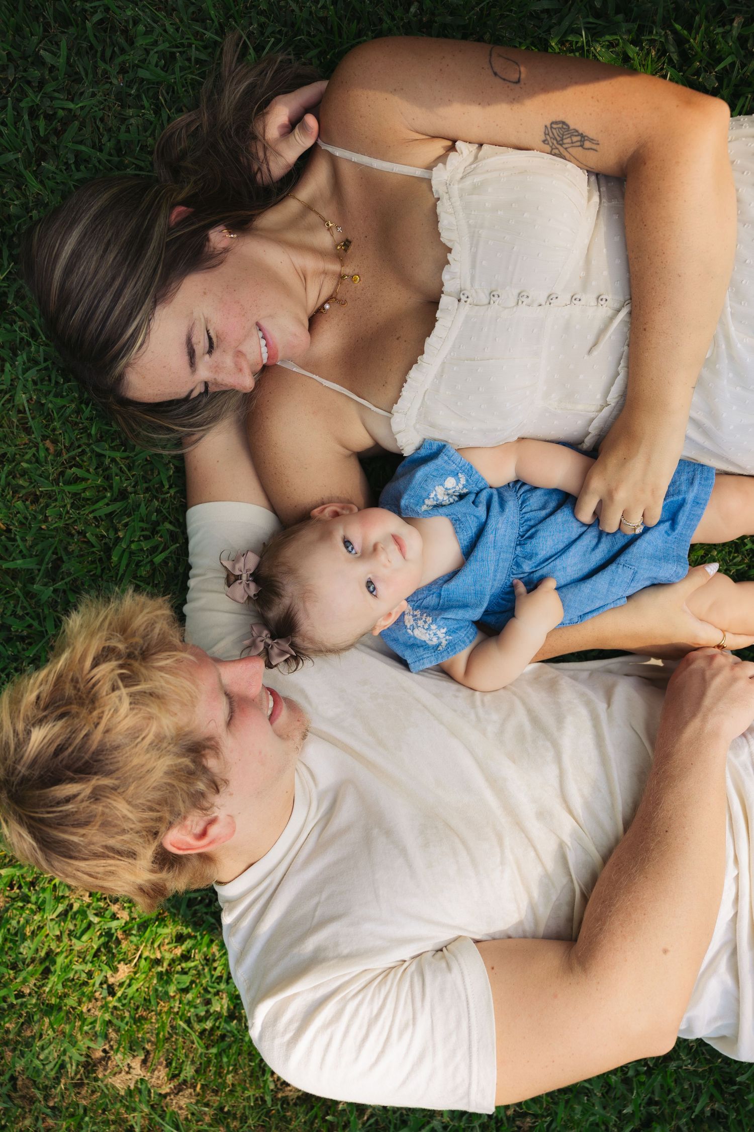 Happy young family lying on grass with their smiling baby in blue outfit.