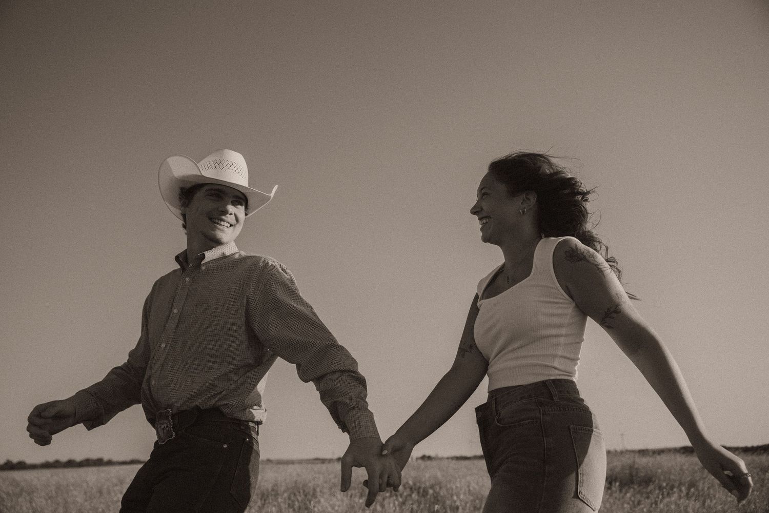 A couple holds hands and runs through a field, the man wearing a cowboy hat, in a romantic sepia-toned outdoor photo.