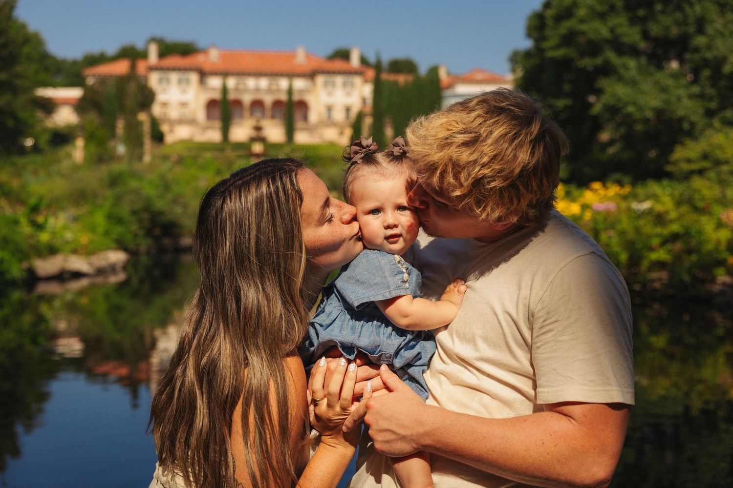 Happy young family with baby kissing outdoors near elegant estate and pond on sunny day.