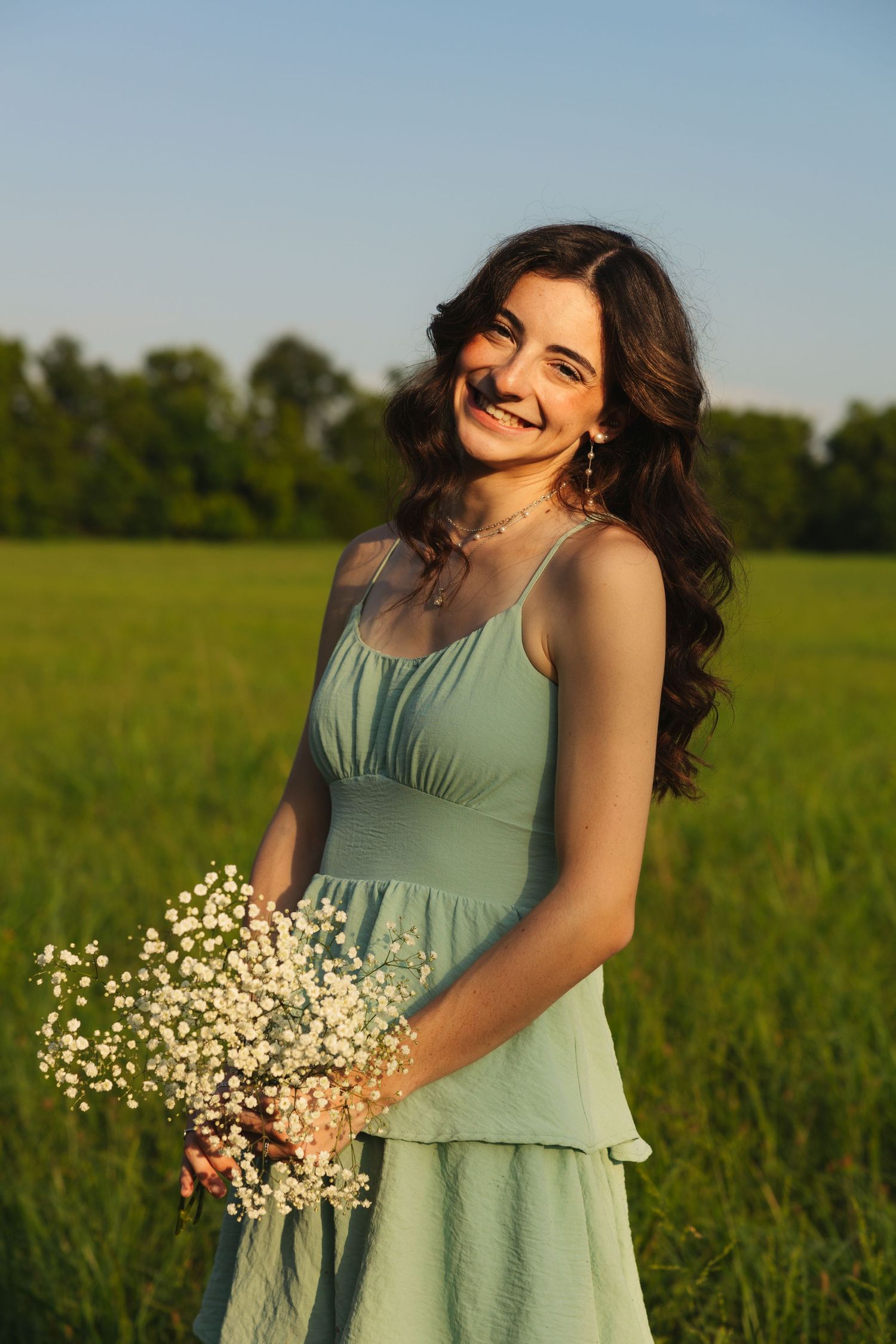 Smiling young woman in mint green dress holding white wildflowers in a sunny green field at sunset.