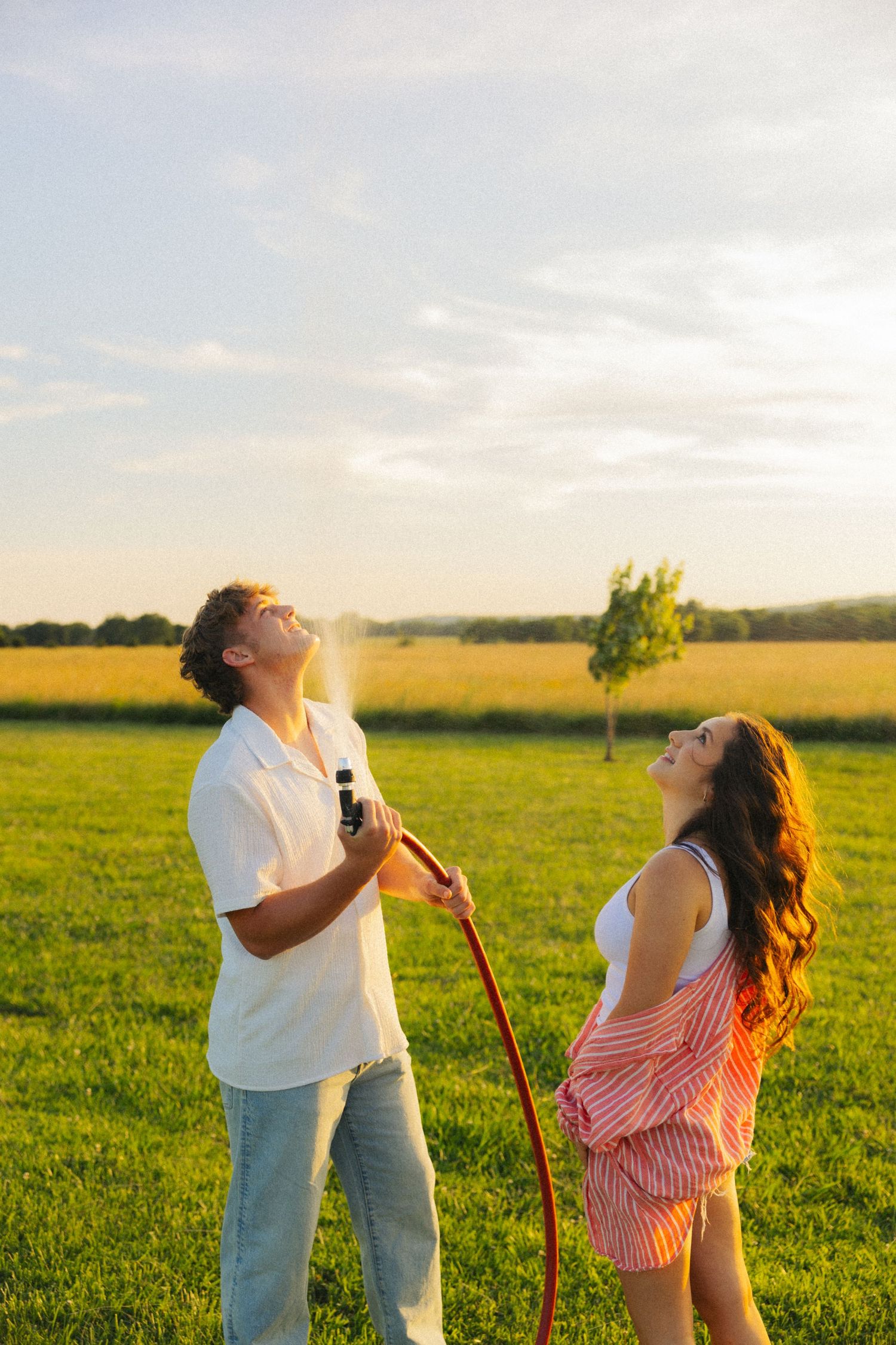 A young couple plays with a garden hose in a sunny green field at sunset, laughing as water sprays into the air.