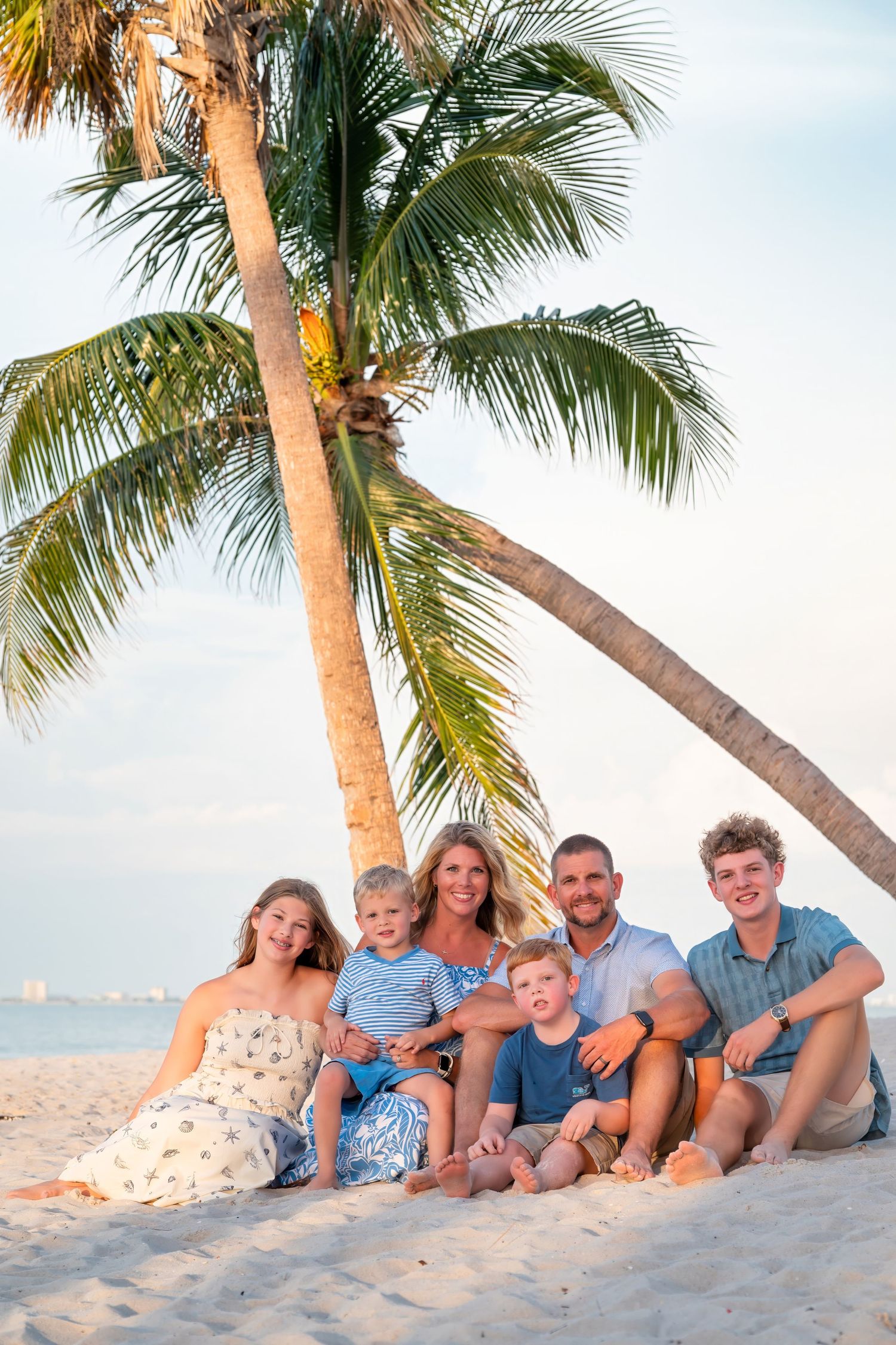 Family of six posing together on a sandy beach under palm trees at sunset in a tropical location.