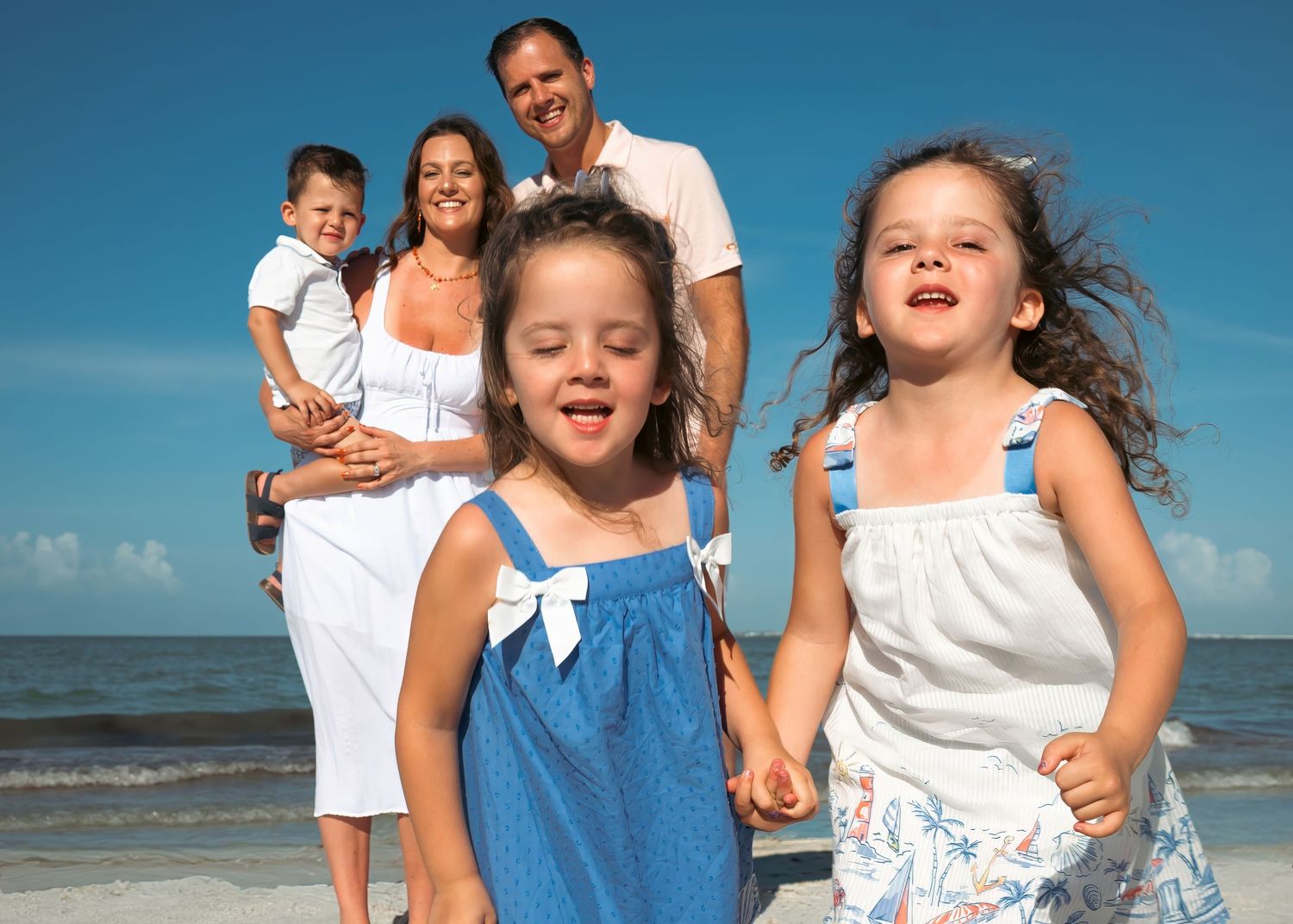 Family of four enjoying a sunny beach day, with two young girls holding hands and parents smiling in the background.