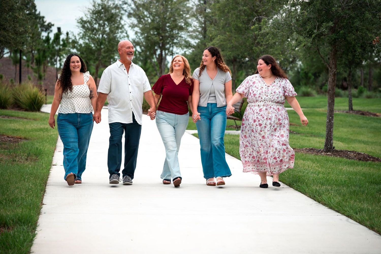 Five family members walk hand-in-hand on a tree-lined path, smiling outdoors on a sunny day.
