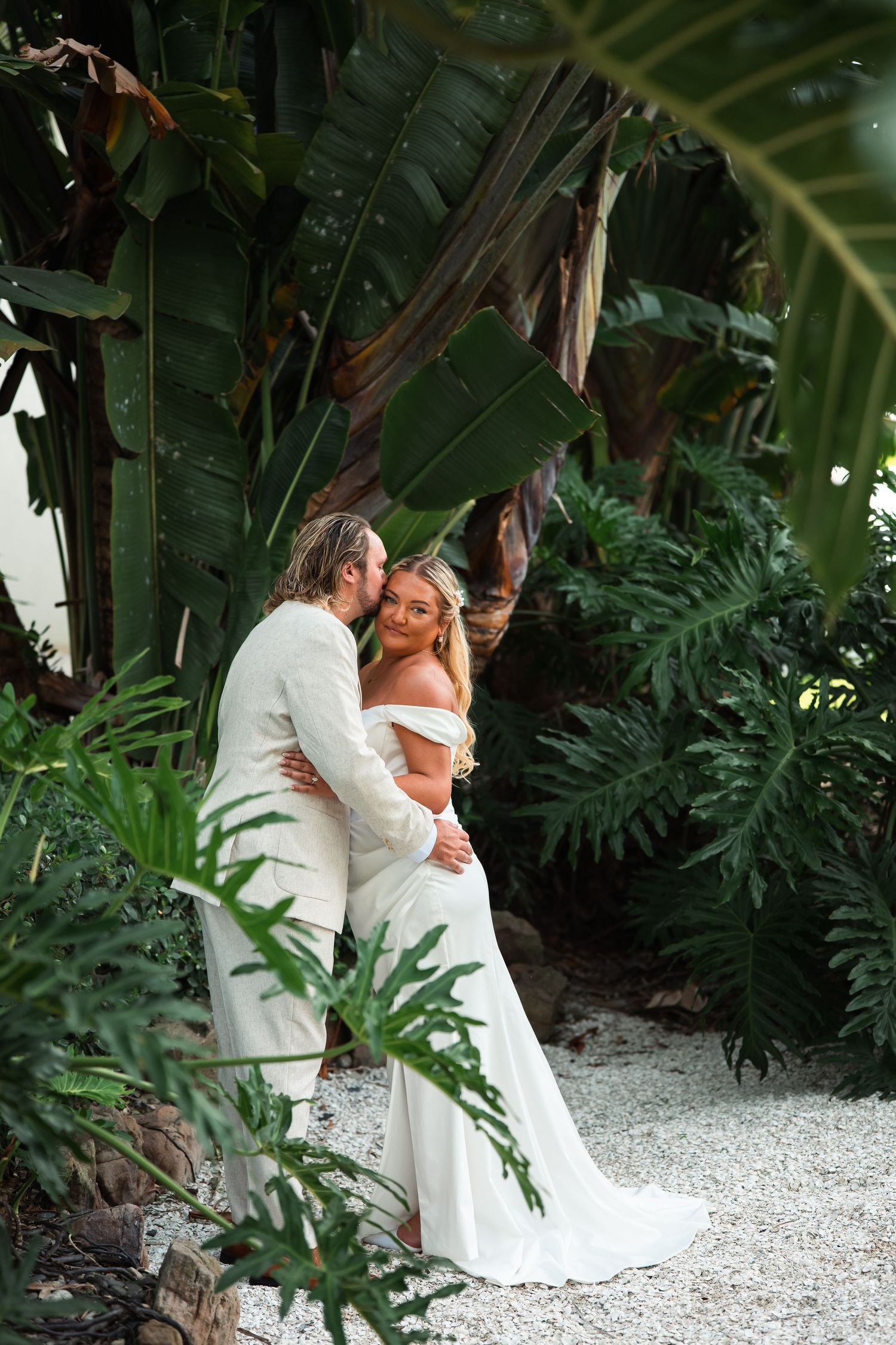 Bride and groom embrace among lush tropical greenery, she in a white off-shoulder gown, he in a cream suit.