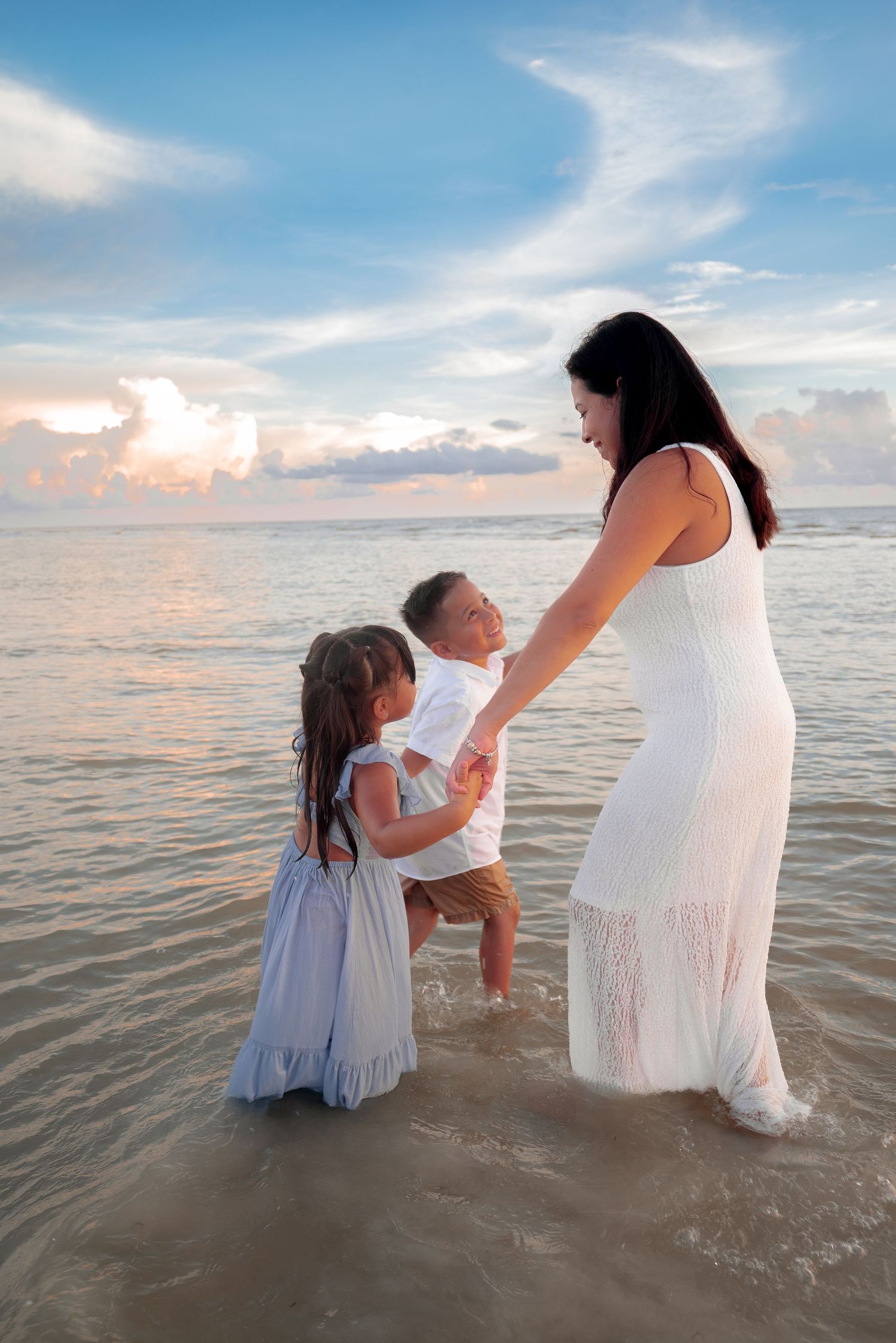 A mother in a white dress stands in ocean waves with her two young children at a beautiful sunset beach.