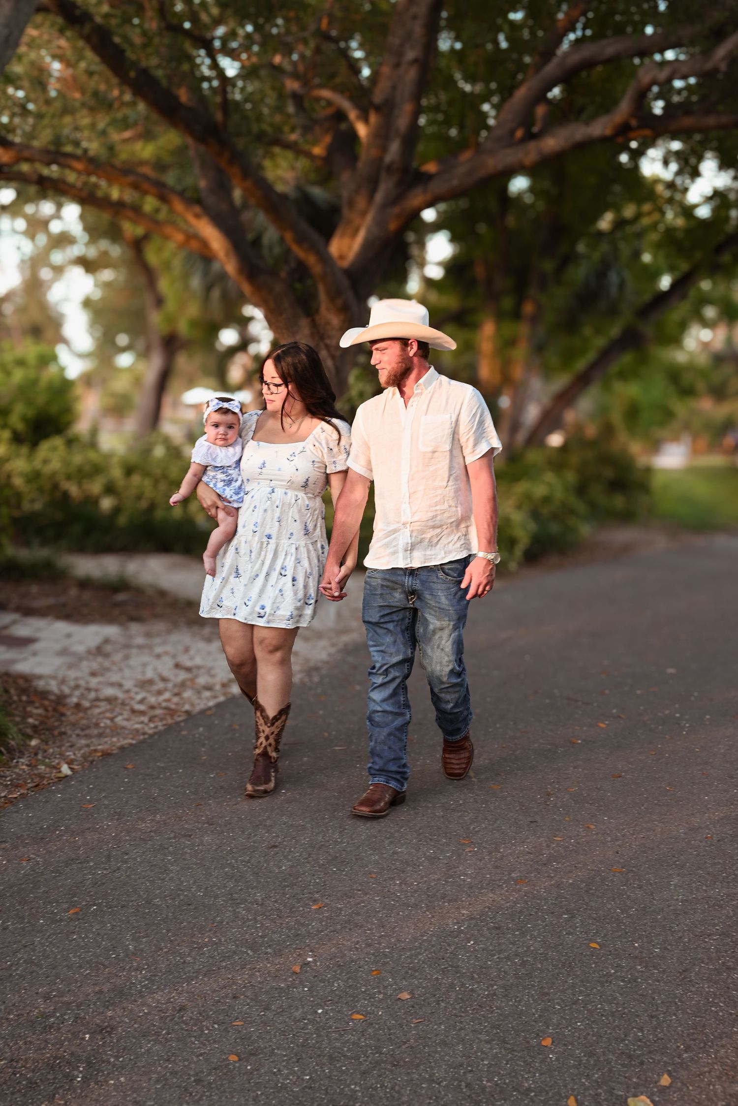 A couple in cowboy boots walks with their baby on a tree-lined path.