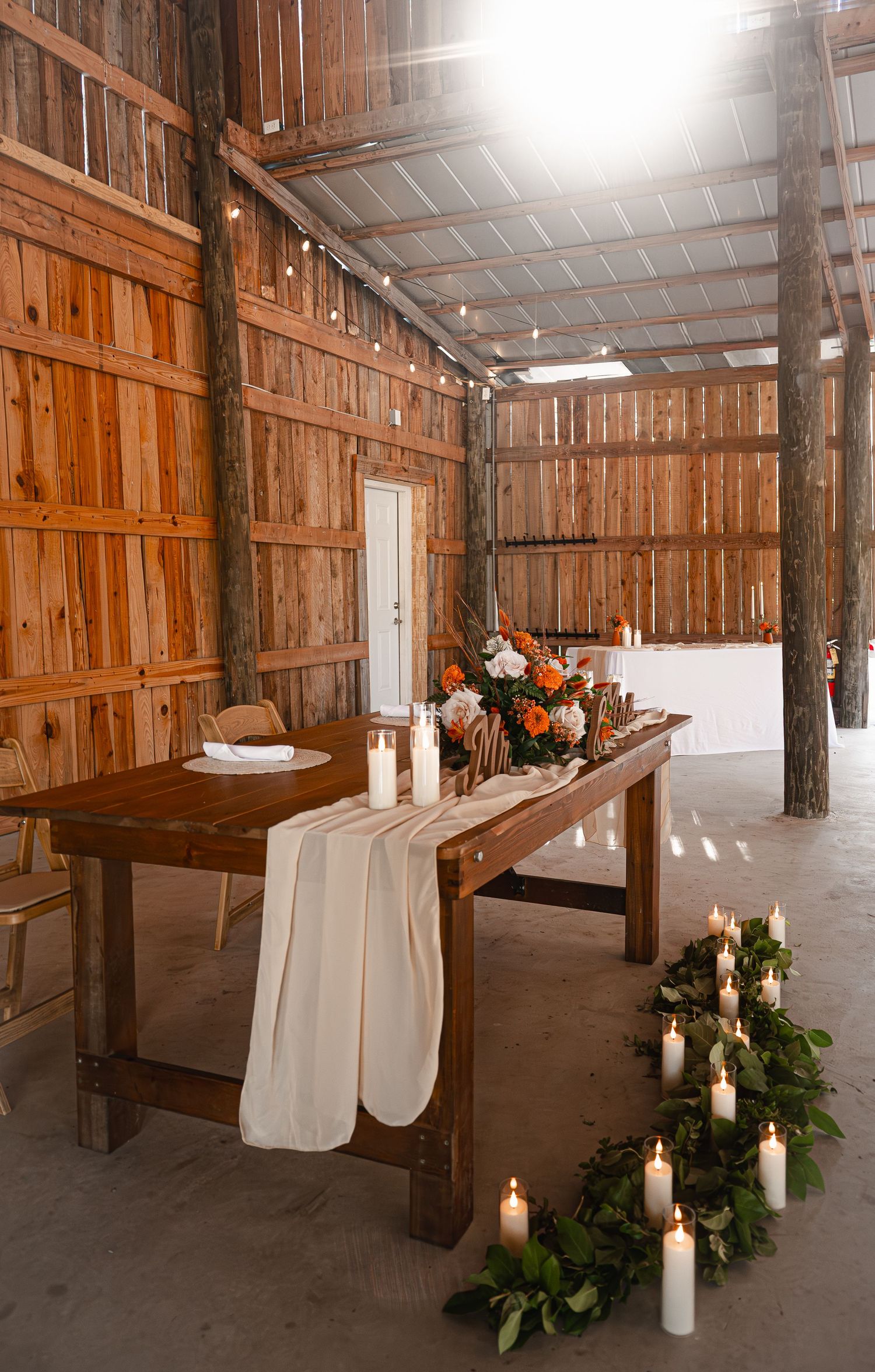 Rustic barn wedding reception table with white linen runner, candles, and floral centerpiece on wood floors.