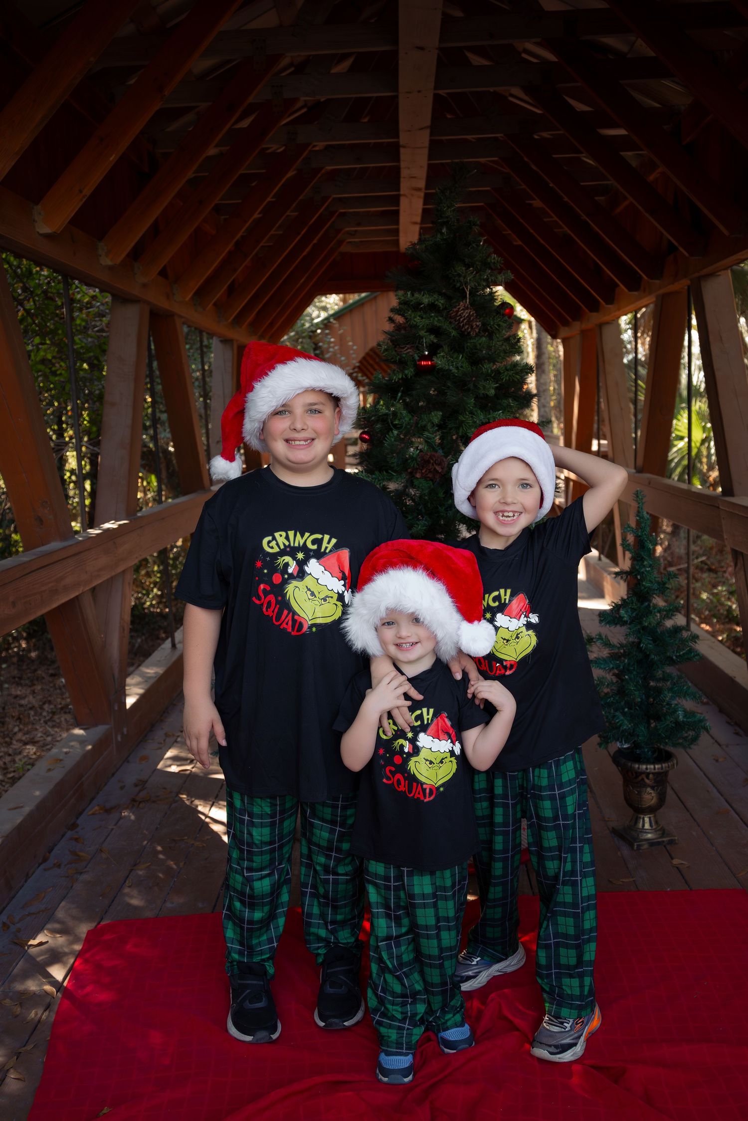 Three children in Santa hats and Grinch Squad shirts pose by a Christmas tree on a wooden bridge.