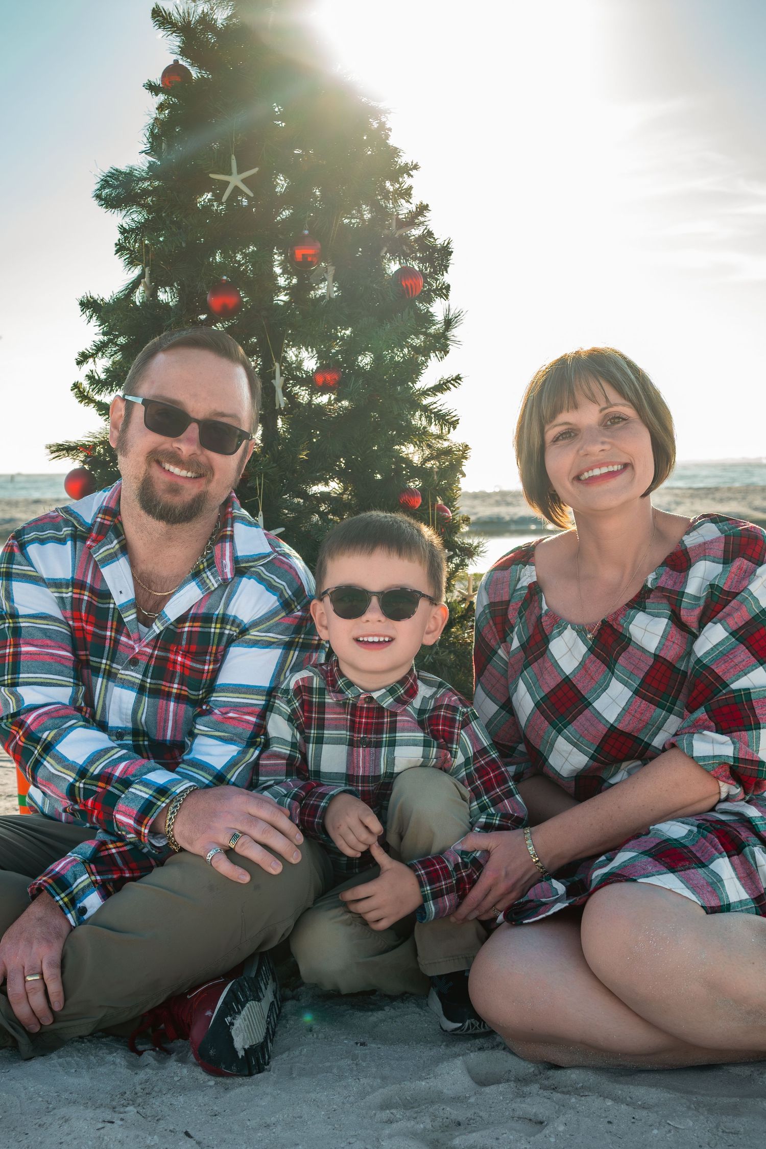 Family of three in matching plaid outfits posing by a decorated Christmas tree on a sunny beach.