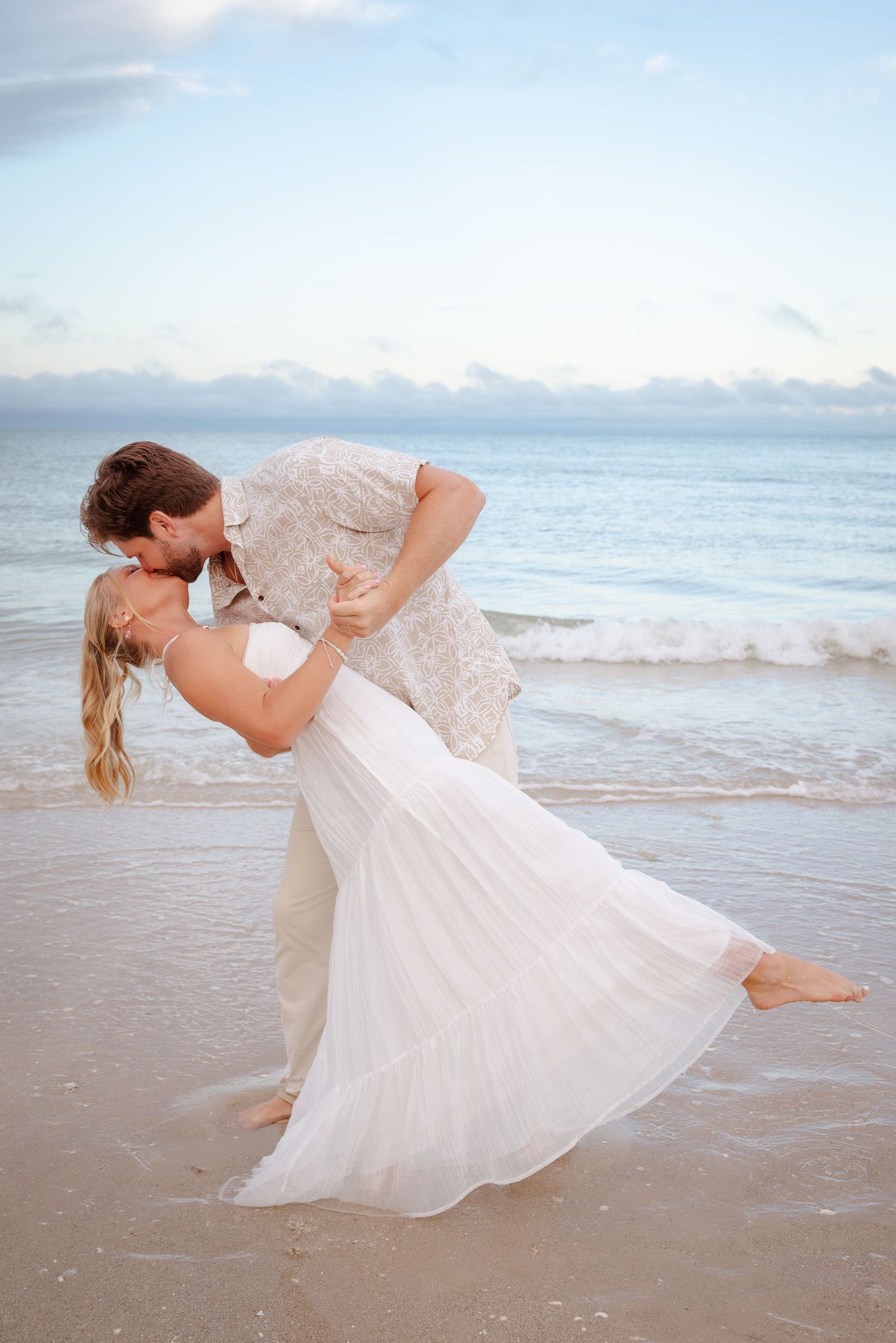 Couple sharing a romantic dip kiss on a sandy beach at sunset, bride in white dress and groom in casual attire.