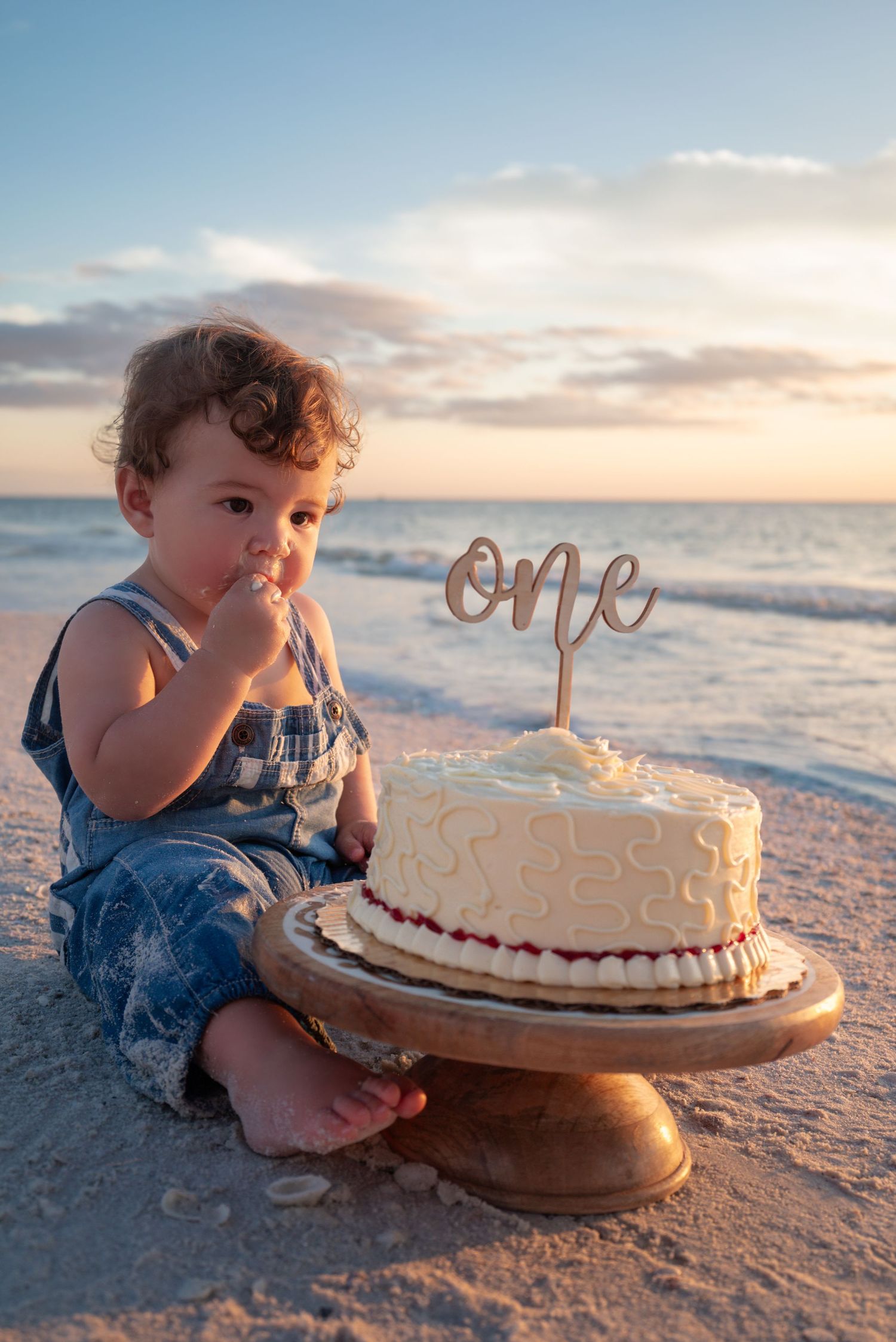 Baby's first birthday cake smash on the beach at sunset with a white cake and 'one' topper.