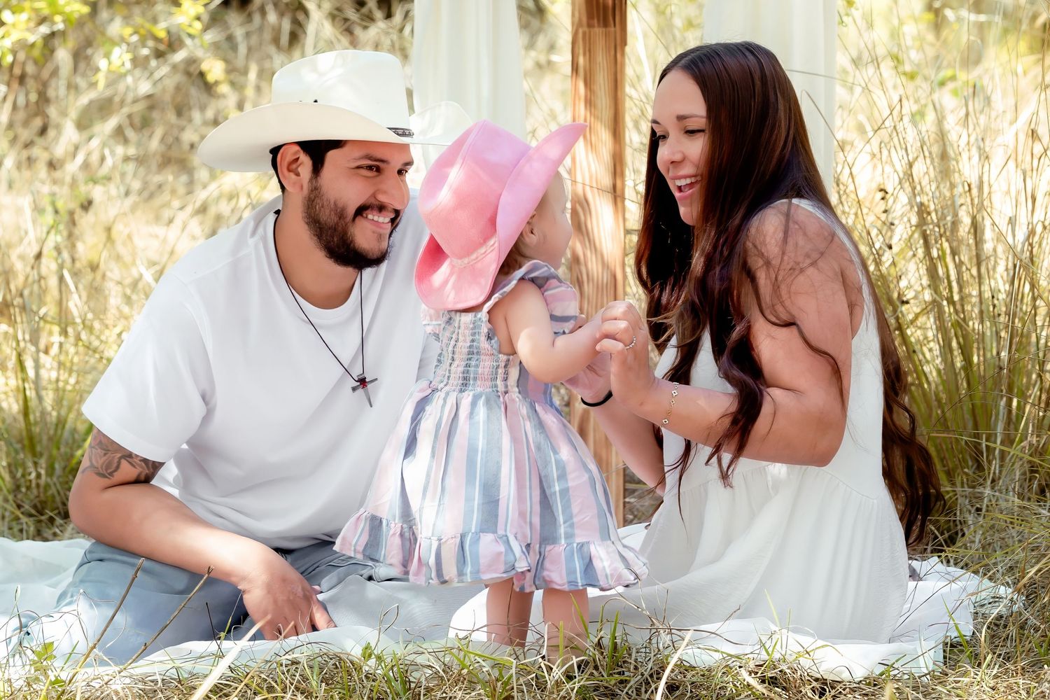 Young couple sitting in grass smiling at their toddler daughter in a pink hat and colorful dress.
