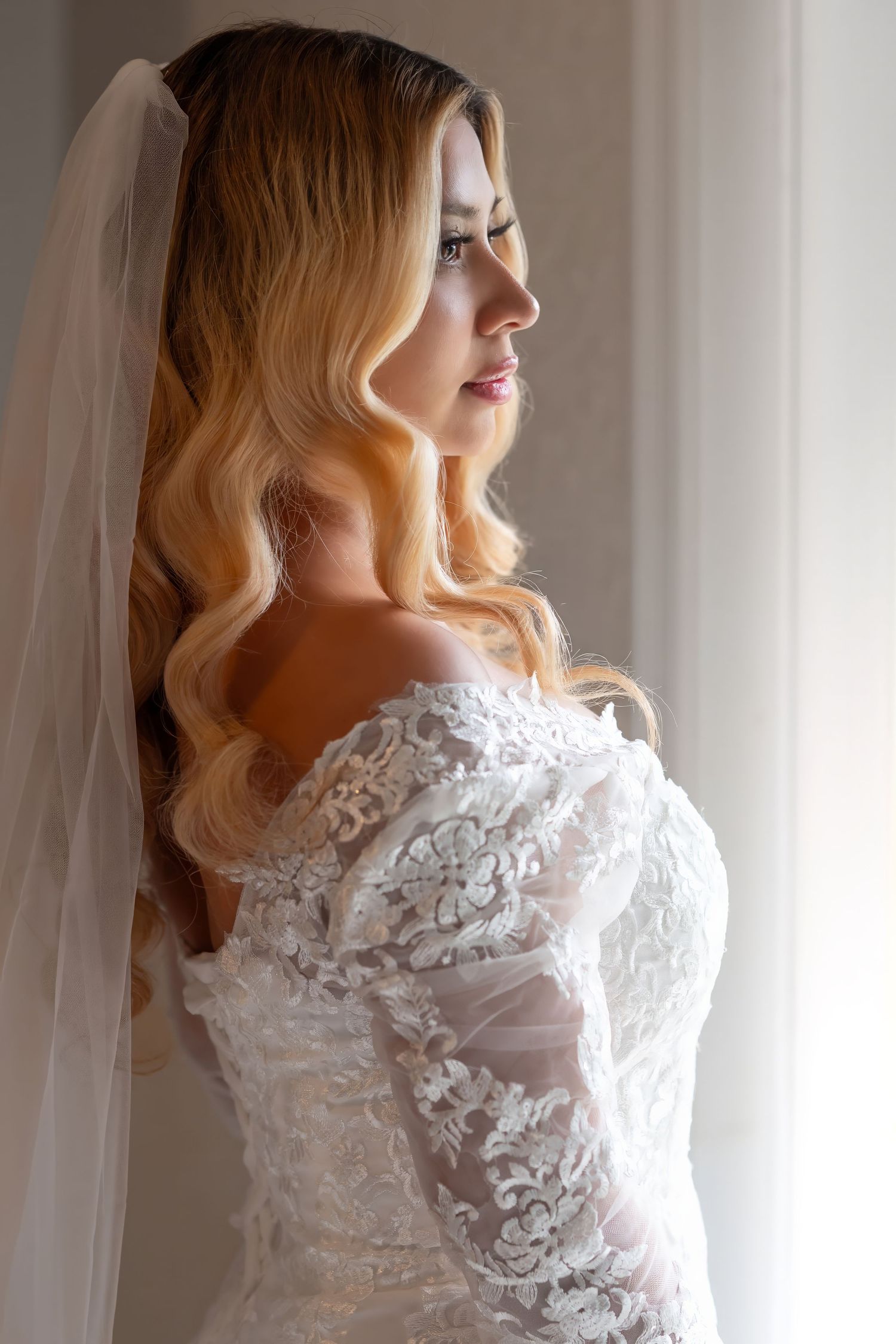 Beautiful blonde bride in off-shoulder lace wedding gown with veil gazing out bright window.