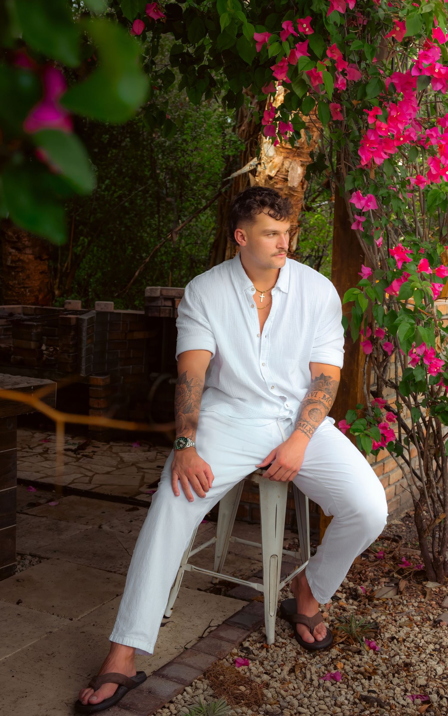 Stylish tattooed man in all-white outfit sits on a stool surrounded by blooming pink bougainvillea flowers.