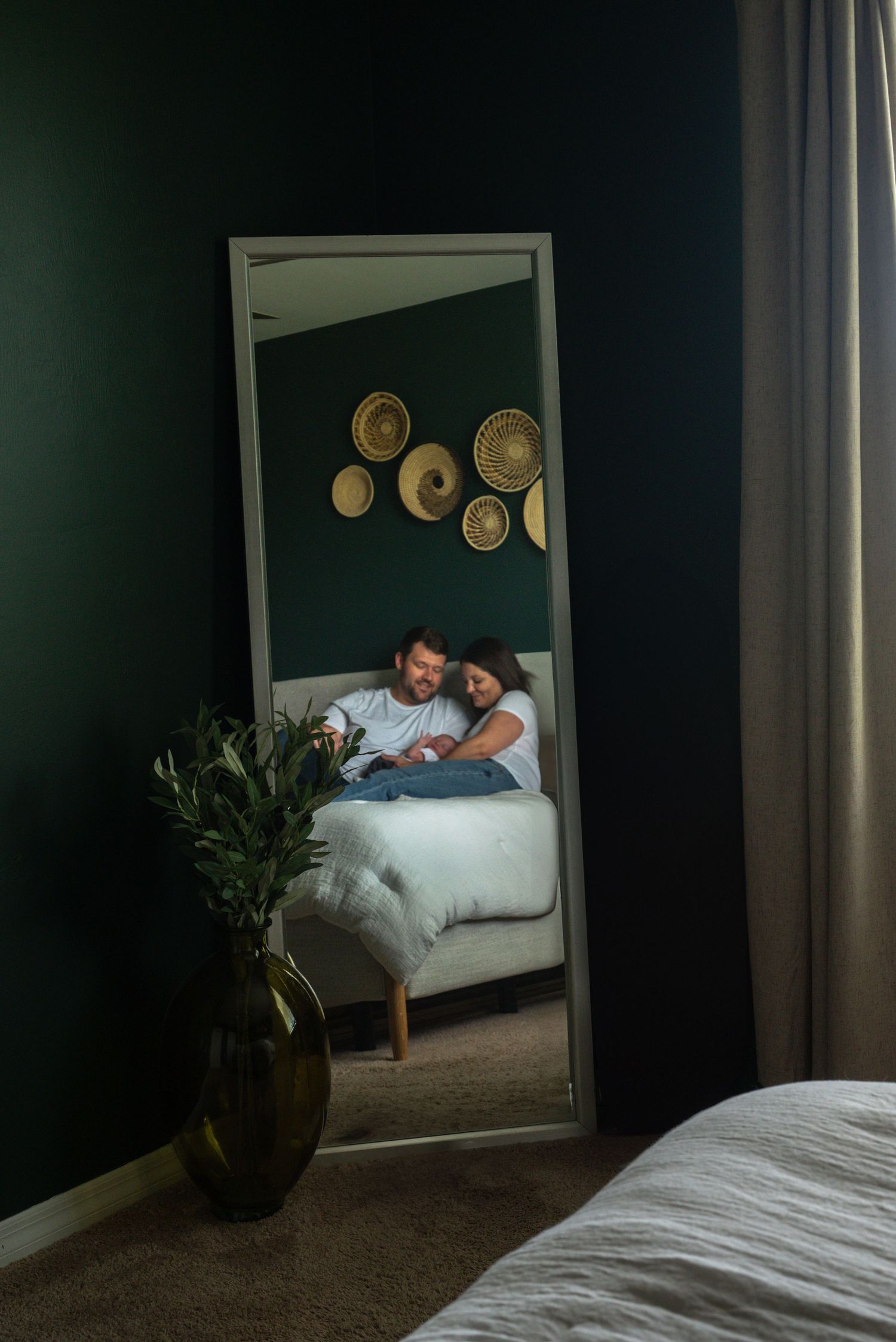 Mirror reflection of couple cuddling on bed in a dark green bedroom with woven wall decor.