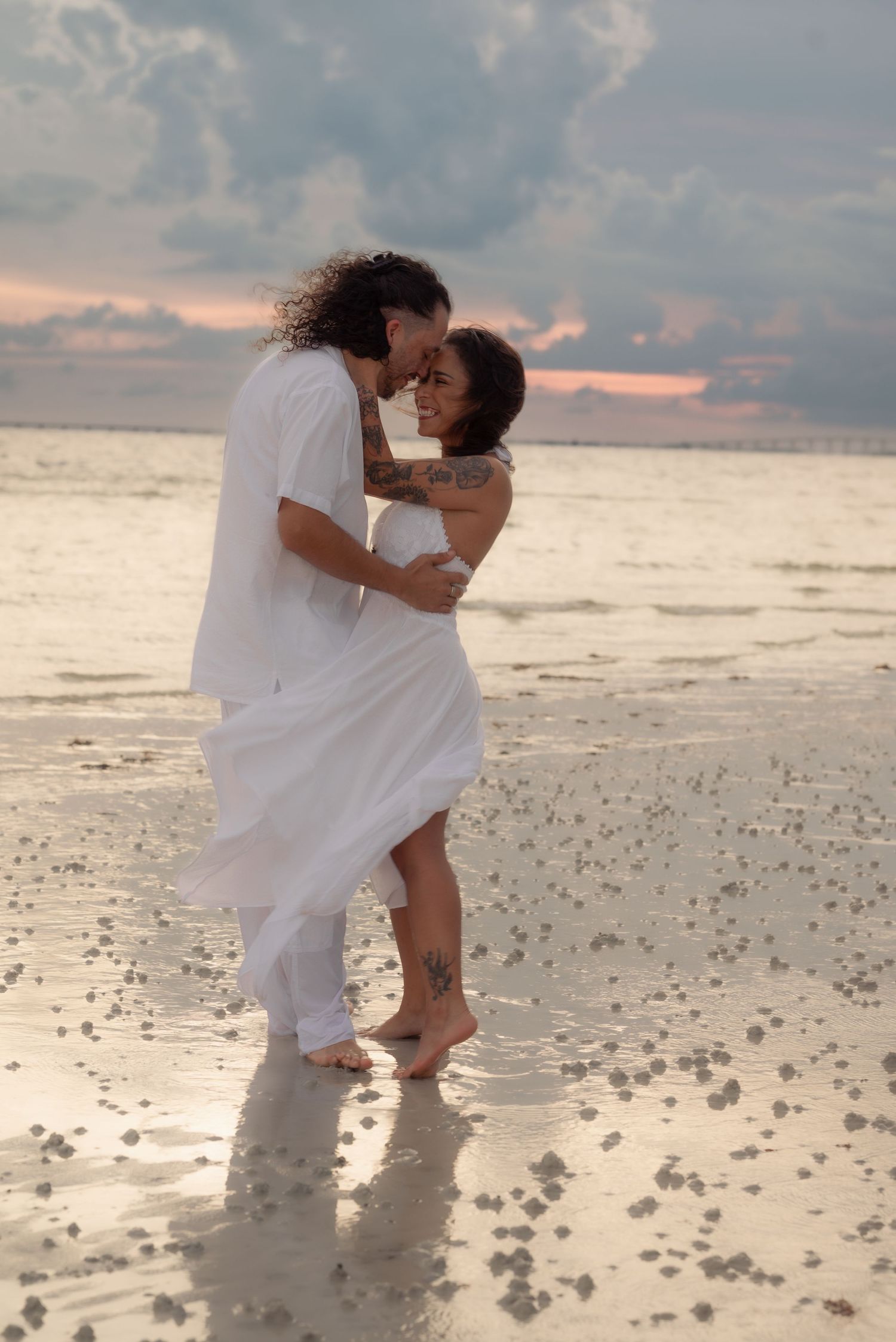 Tattooed couple in white embracing barefoot on wet sandy beach at sunset with cloudy sky.