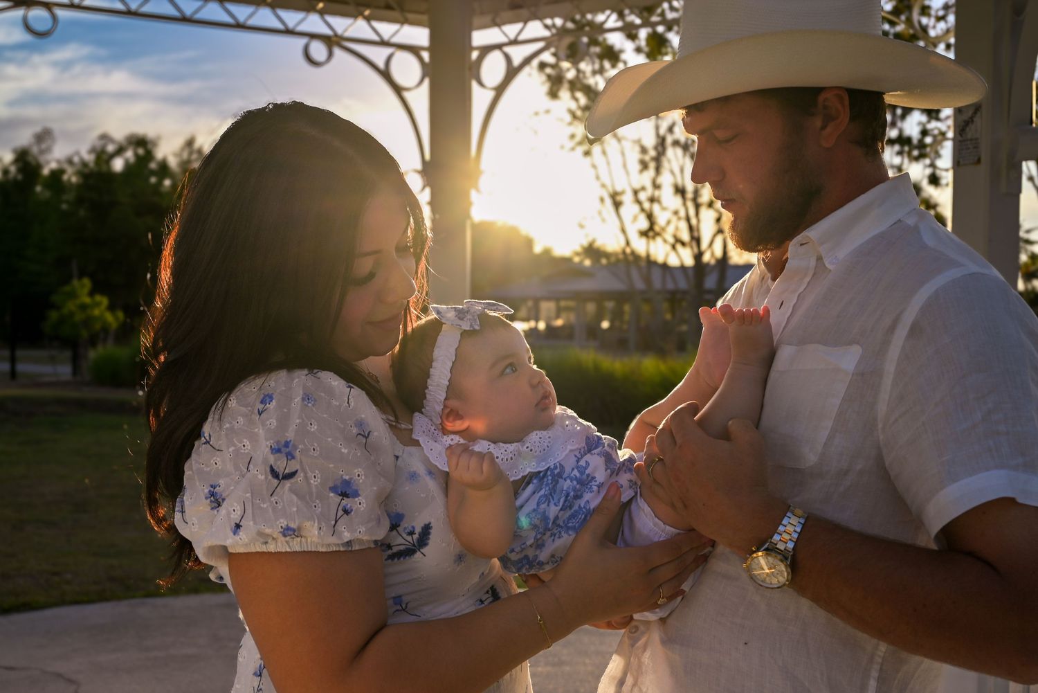 Young family with baby at golden hour sunset in park, parents smiling at newborn in floral outfit.