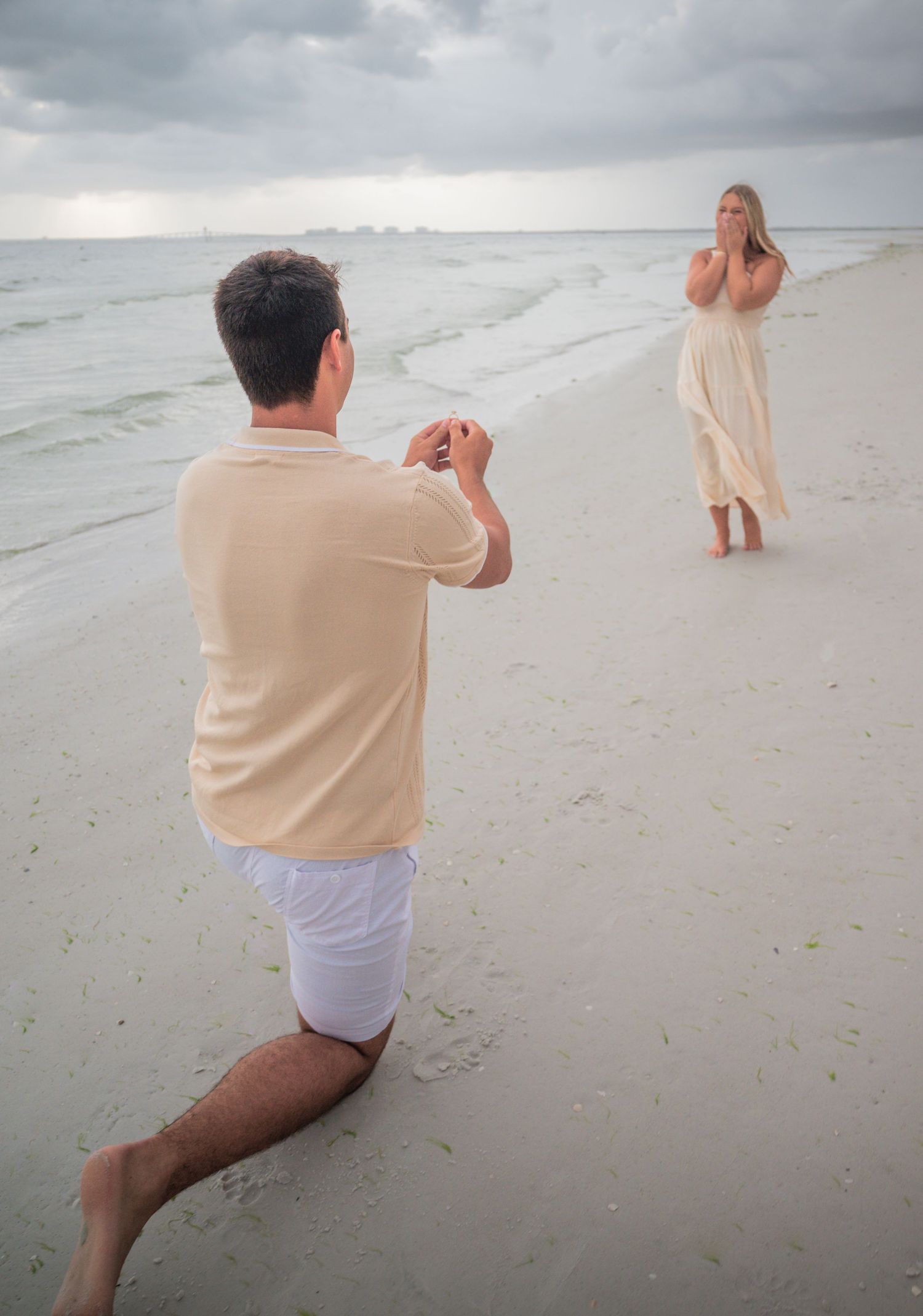 Man proposing on one knee to surprised woman on a sandy beach at dusk.