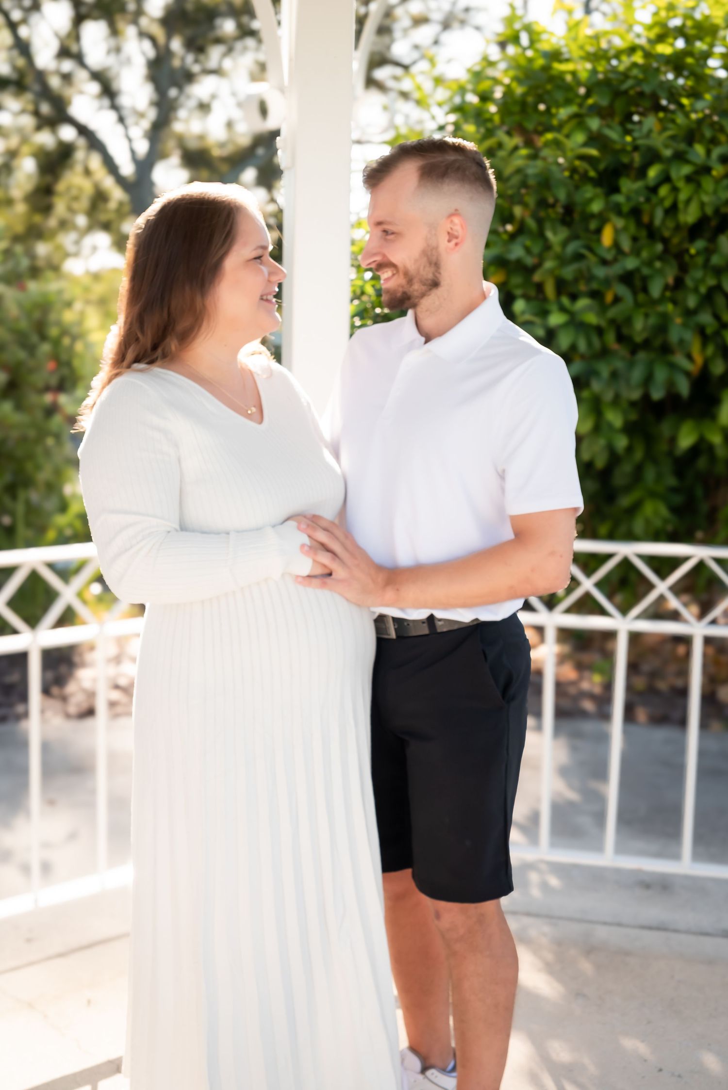 A smiling pregnant couple in white outfits standing together on a sunny porch.
