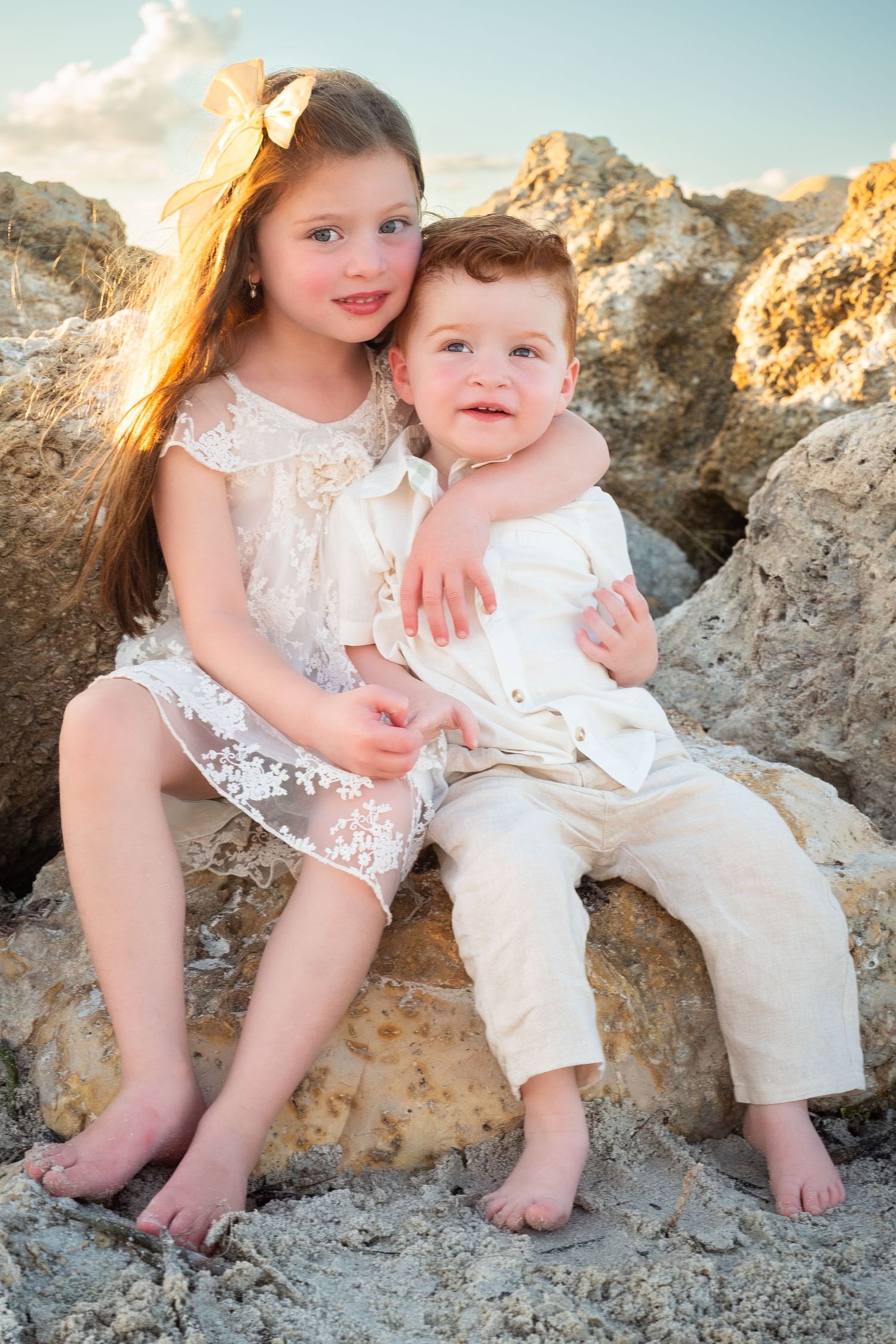 Sweet siblings in white outfits sitting together on rocky beach, older sister hugging younger brother.
