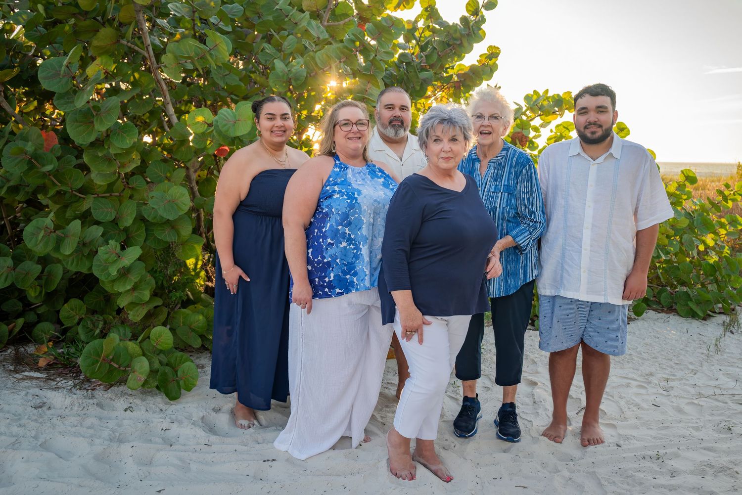Group of six adults posing together on sandy beach surrounded by lush green tropical foliage.