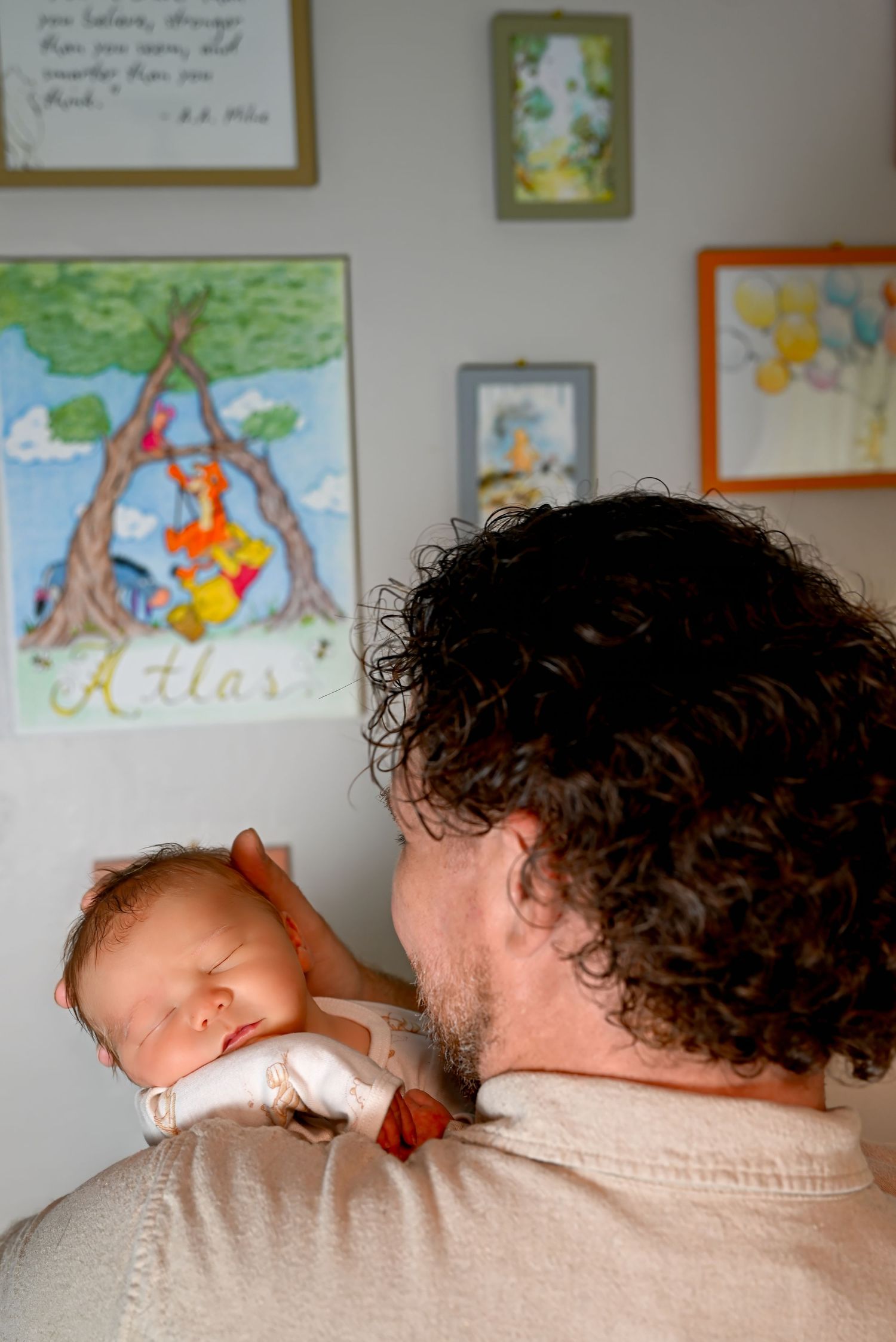 Father with curly hair holds sleeping newborn baby in nursery decorated with Winnie the Pooh art and name Atlas.