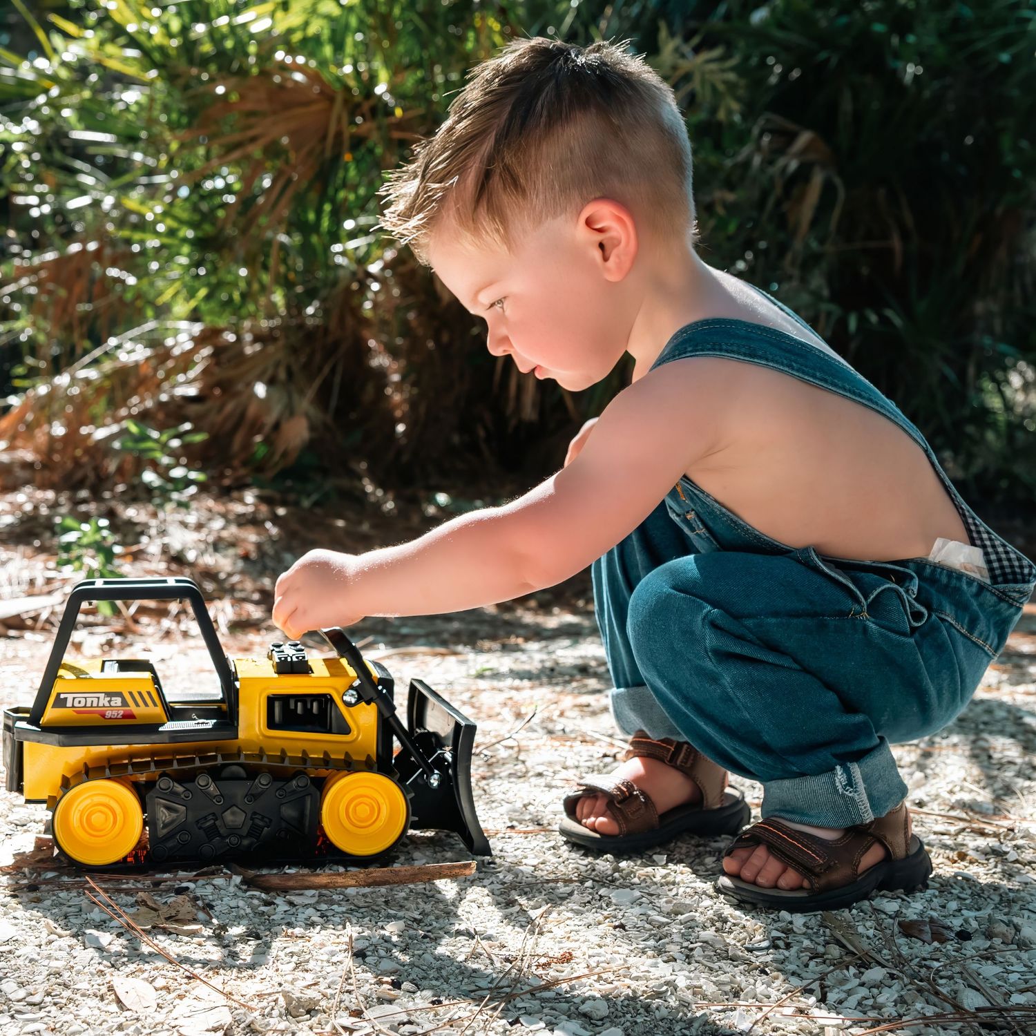 Young boy in denim overalls playing with a yellow toy construction bulldozer outdoors on sandy ground.