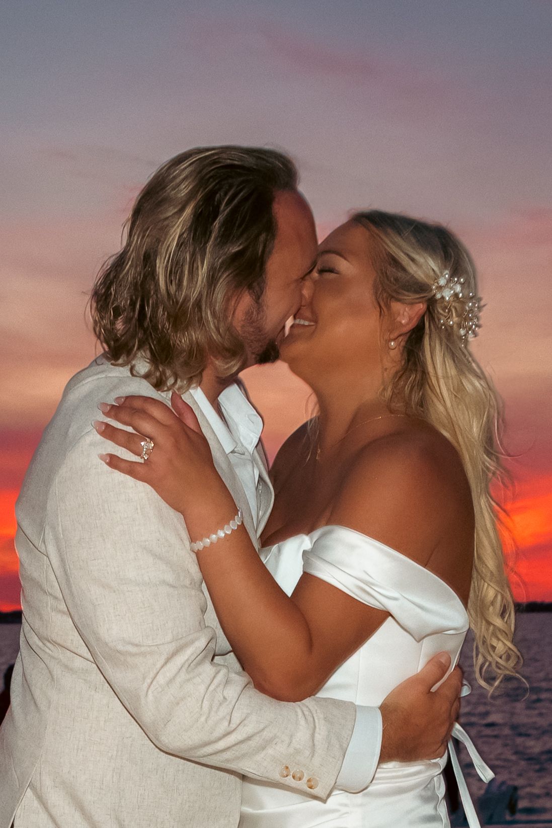 Bride and groom sharing a romantic kiss at sunset by the water, bride in white off-shoulder gown with floral hair accessory.