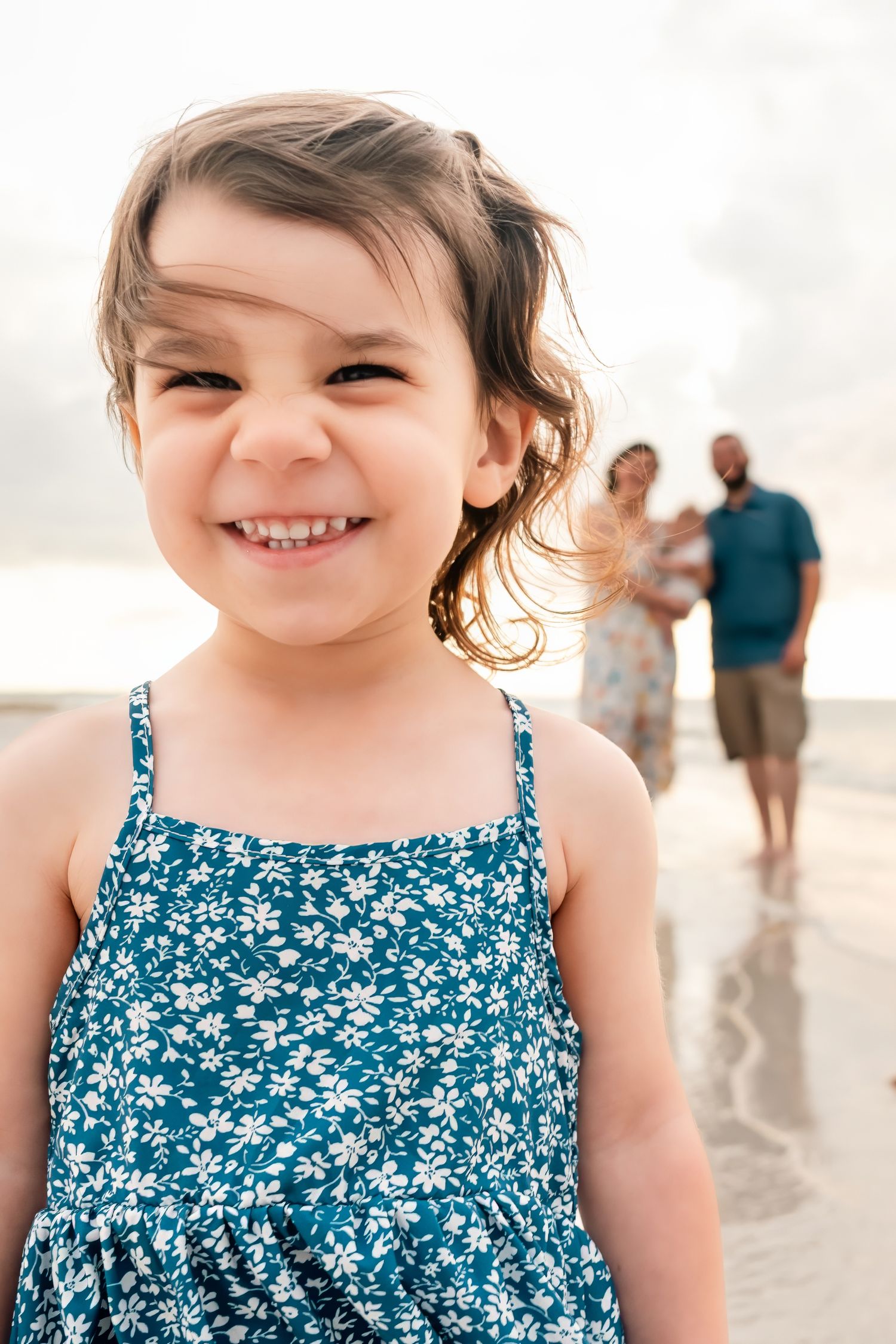 A smiling young girl in a floral blue dress on a beach with her family in the background.