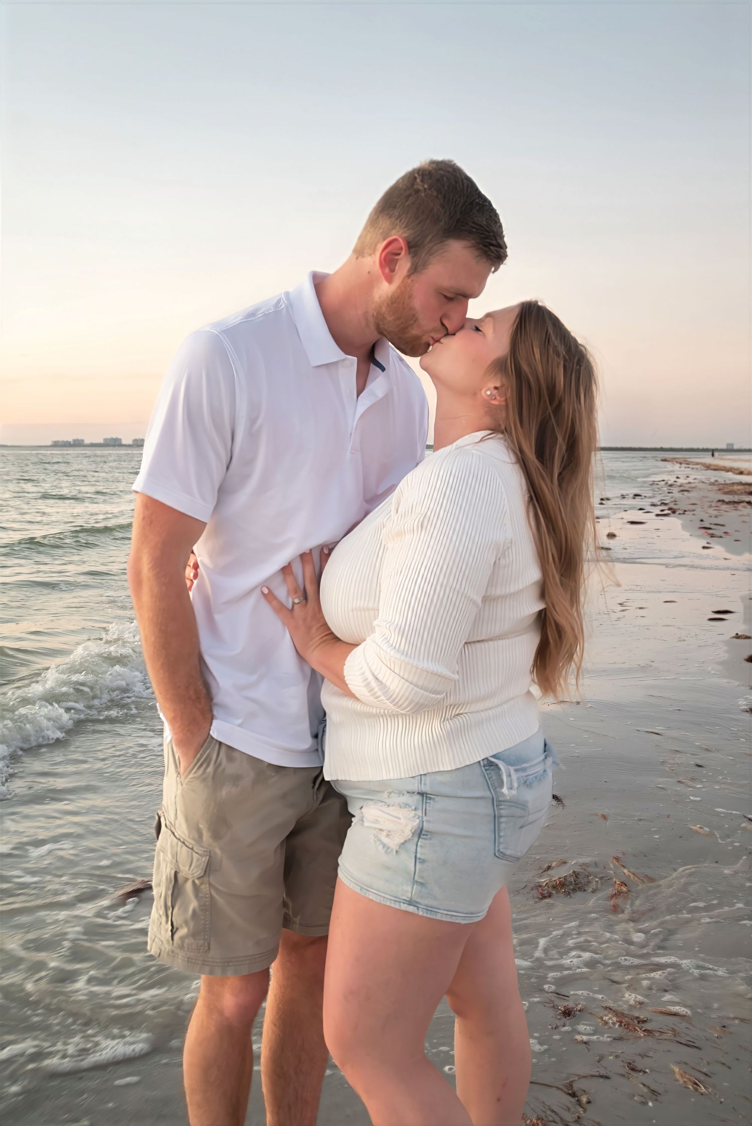 Couple sharing a romantic kiss on the beach at sunset, wearing white tops and casual shorts.